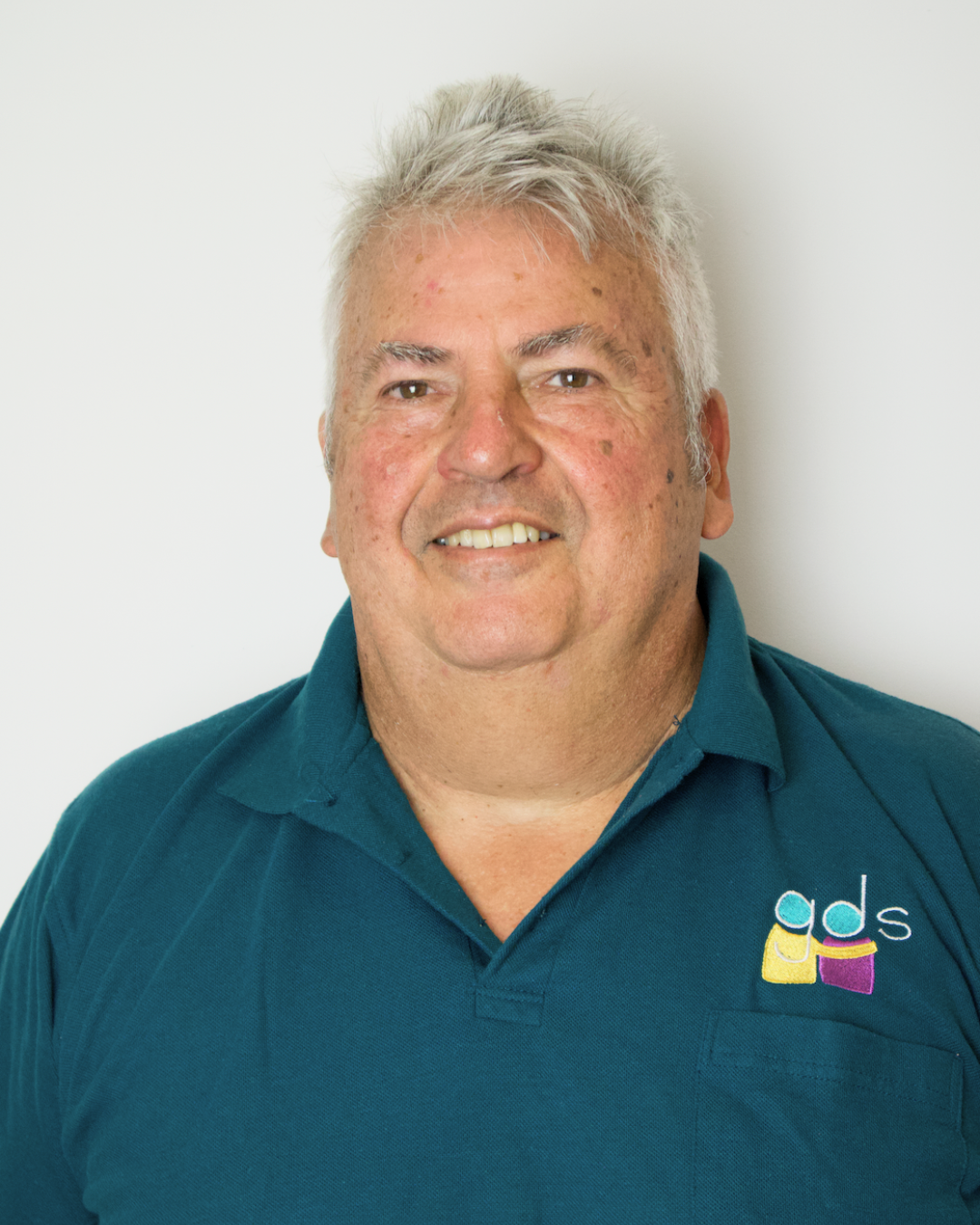 A middle-aged man with gray hair, smiling, wearing a teal polo shirt with a logo that reads 'gds' and features colorful abstract shapes.