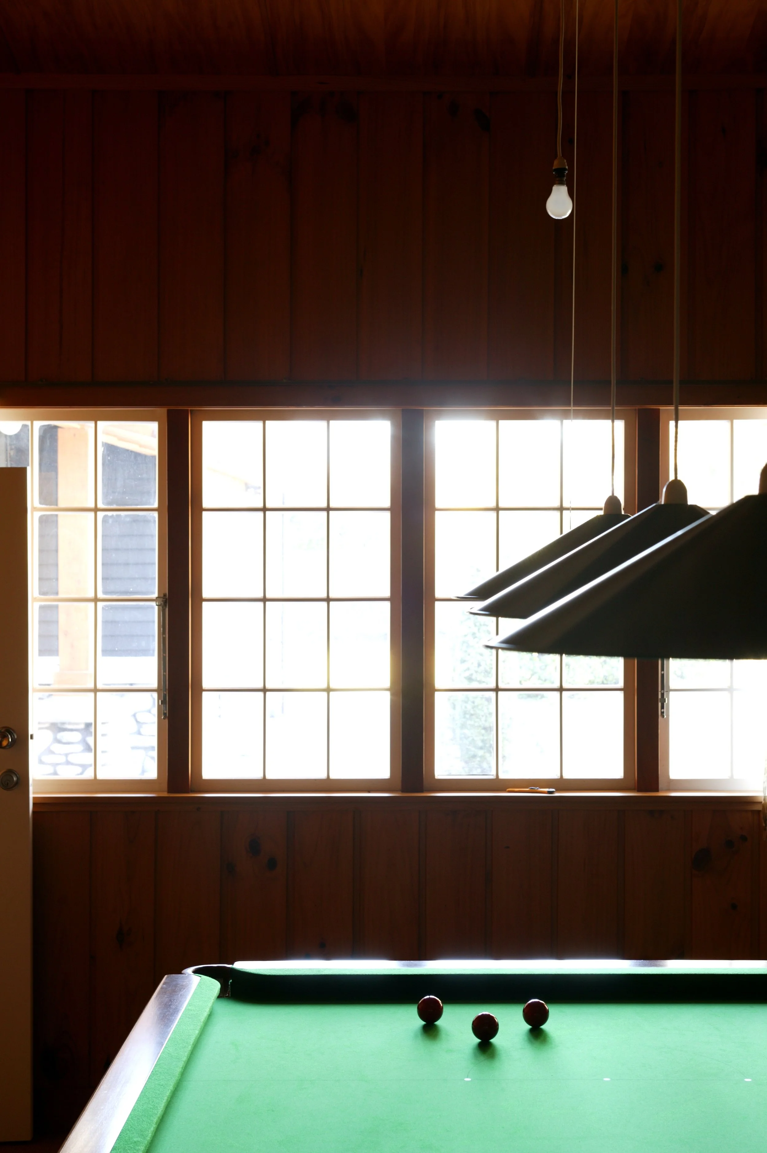 Interior of a wooden game room with a pool table, black pendant lights, large window, and hanging light bulb.