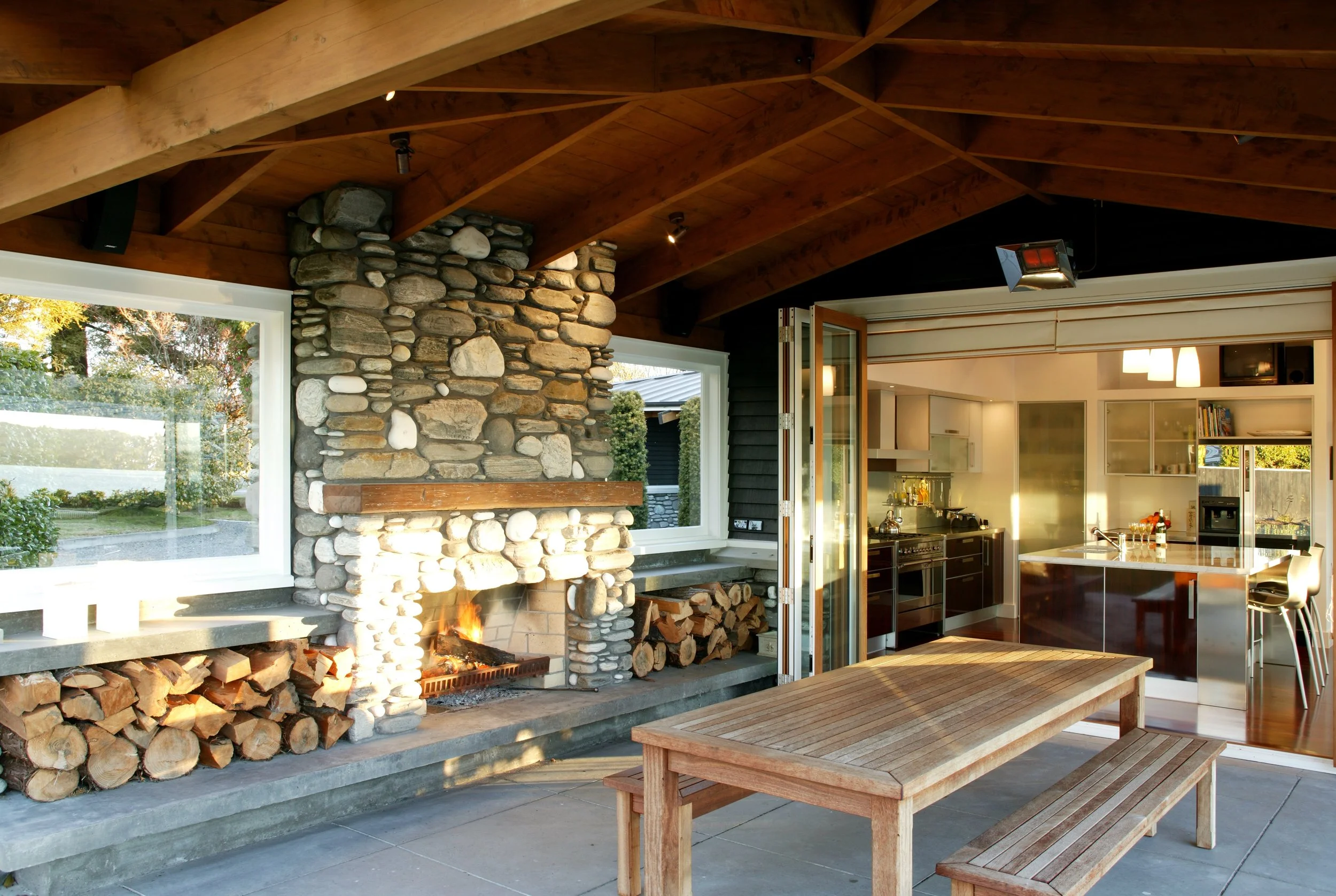 Indoor outdoor kitchen and living area with a stone fireplace, wooden ceiling, and a wooden table with benches.