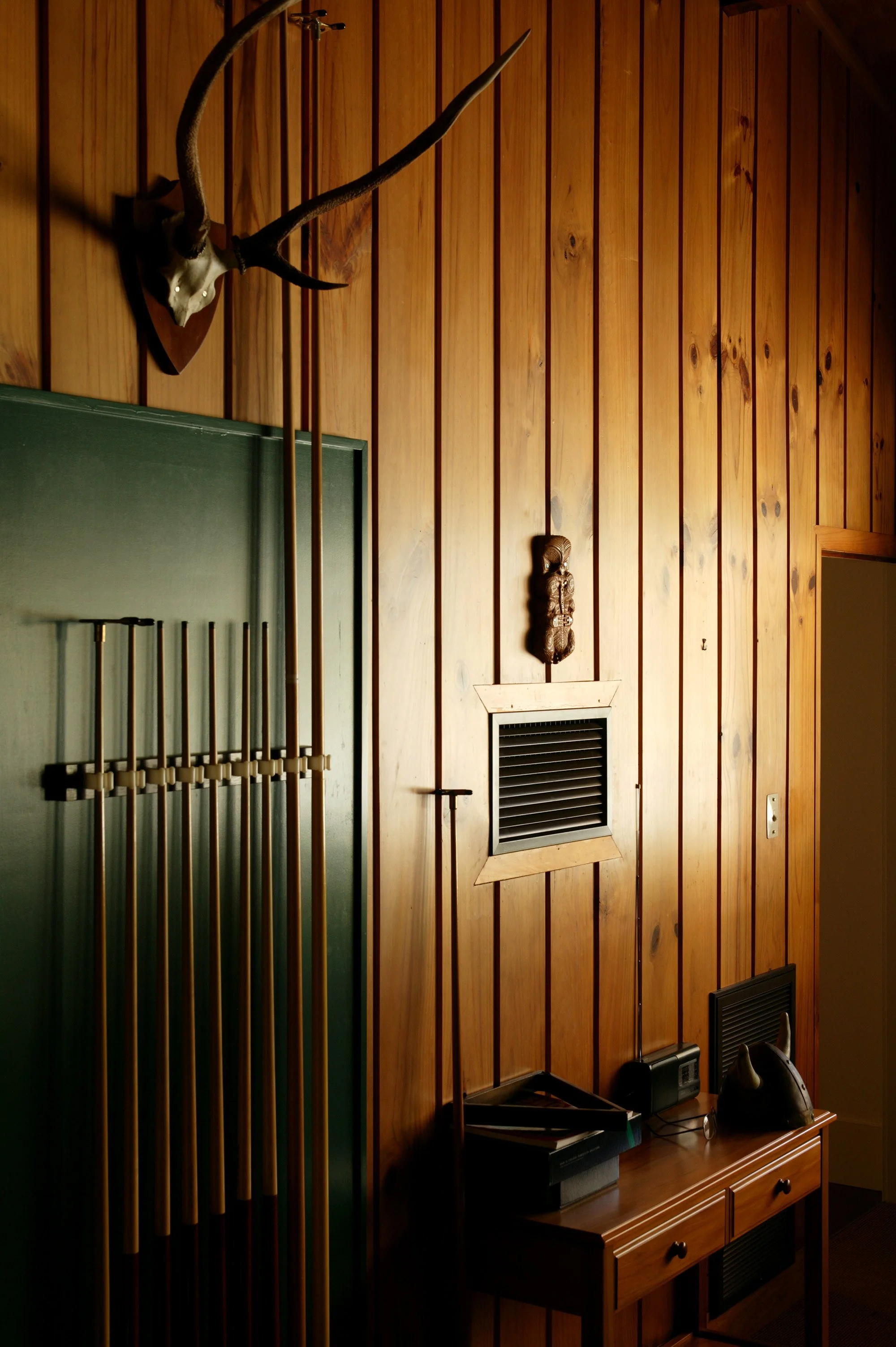 Wood-paneled wall with a mounted deer antelope head, a tribal mask, and a small vent. Below, a wooden table holds a radio, a helmet, and a few other items.