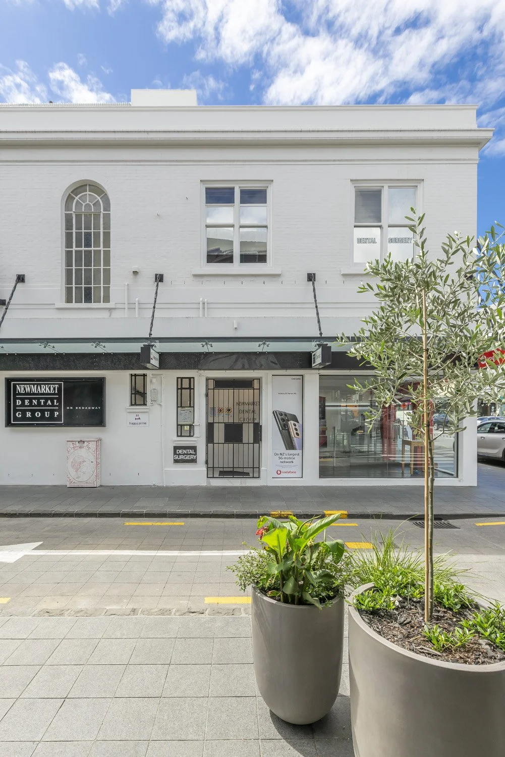 Exterior of a white building housing a dental group, with signage indicating dental services, on a sunny day with a bright blue sky and some scattered clouds. There are two large potted plants in the foreground, a tree and leafy plants.