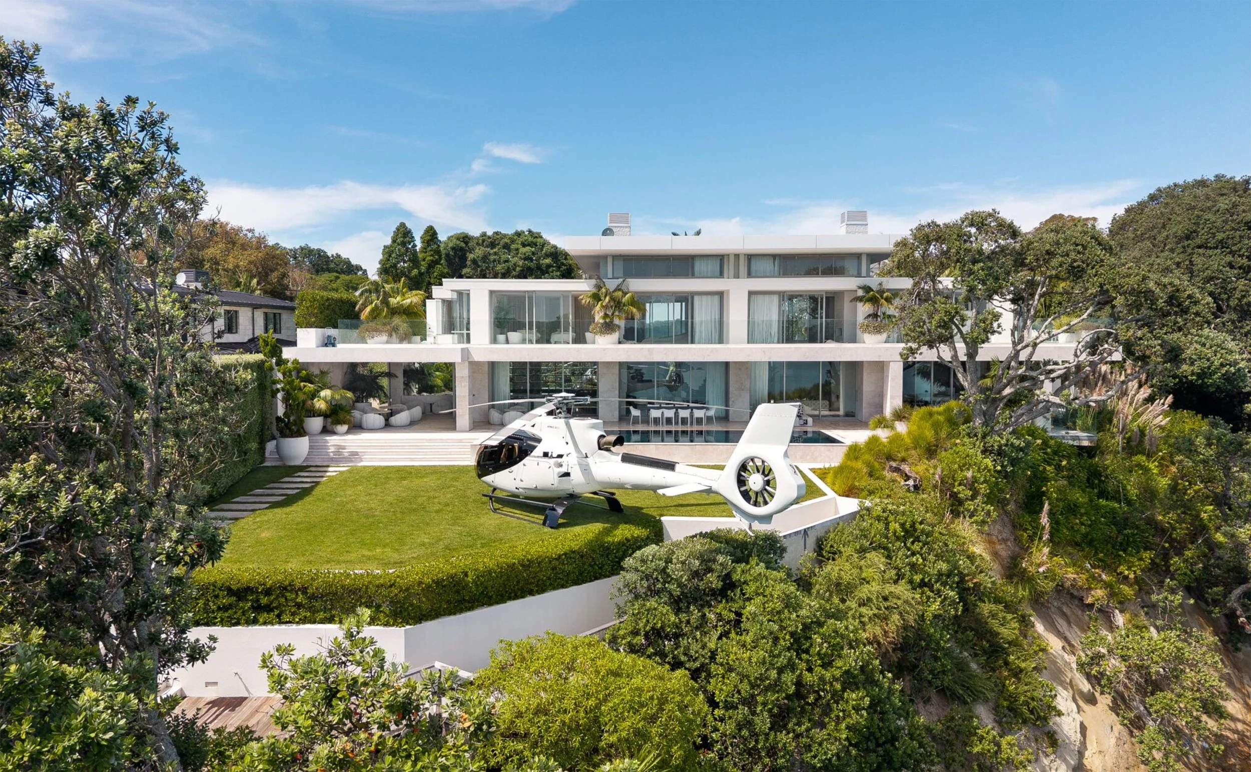 Modern multi-level house with glass walls, surrounded by lush greenery, featuring a white helicopter parked on the lawn in front.