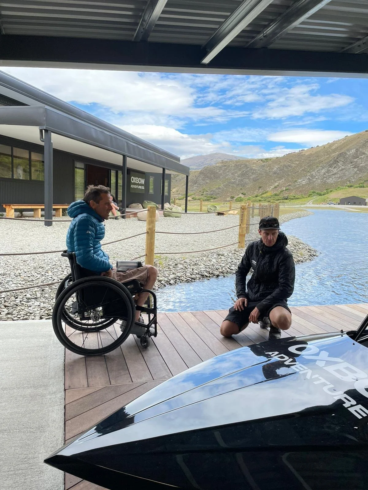 Men sitting outdoors near a body of water with mountains in the background, one in a wheelchair and the other crouching, with a building labeled 'OXBOW ADVENTURE CO.' nearby.