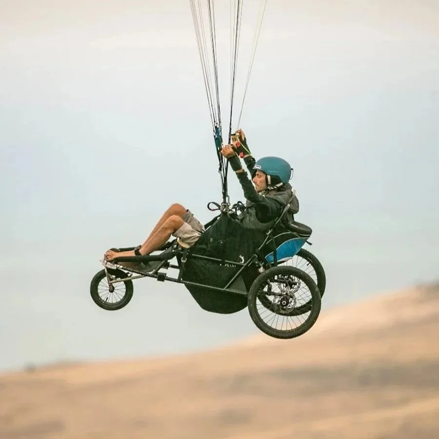 A person in a specialist buggy flying through the air with a parachute, wearing a helmet and gloves.