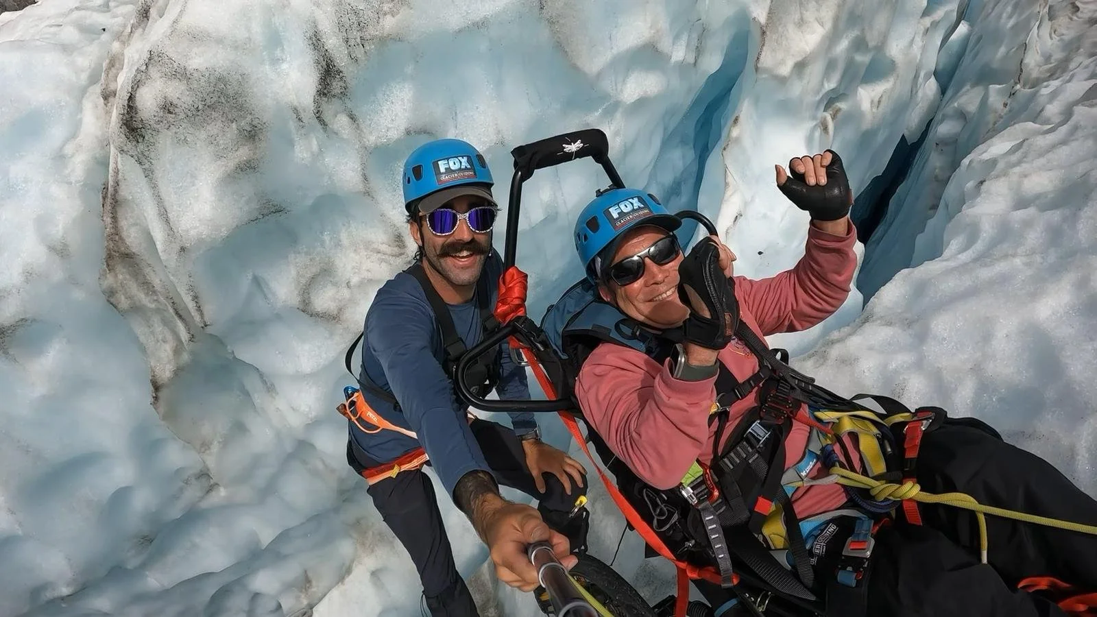 Two men riding a bicycle through a crevasse in the ice, wearing helmets, sunglasses, and cold-weather gear, with one man taking a selfie.