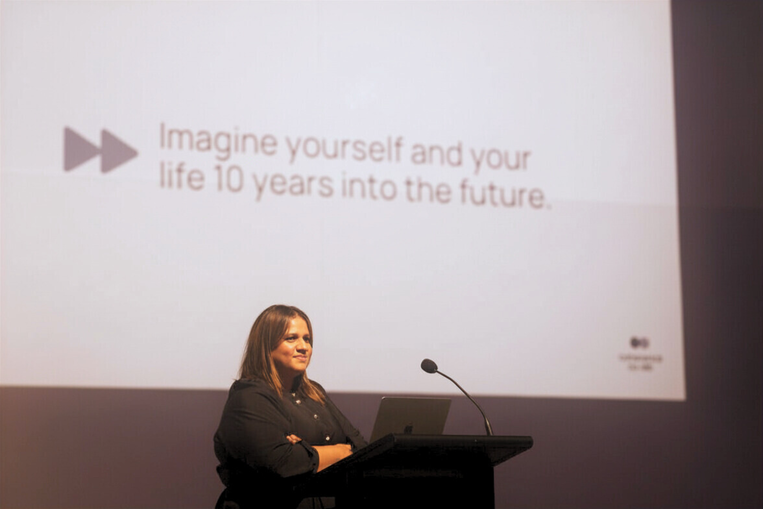 A woman with long brown hair stands at a podium with her arms crossed, looking to her left during a presentation in a conference room. Behind her, a large screen displays text that reads: 'Imagine yourself and your life 10 years into the future,' with a pause icon on the left side of the text.