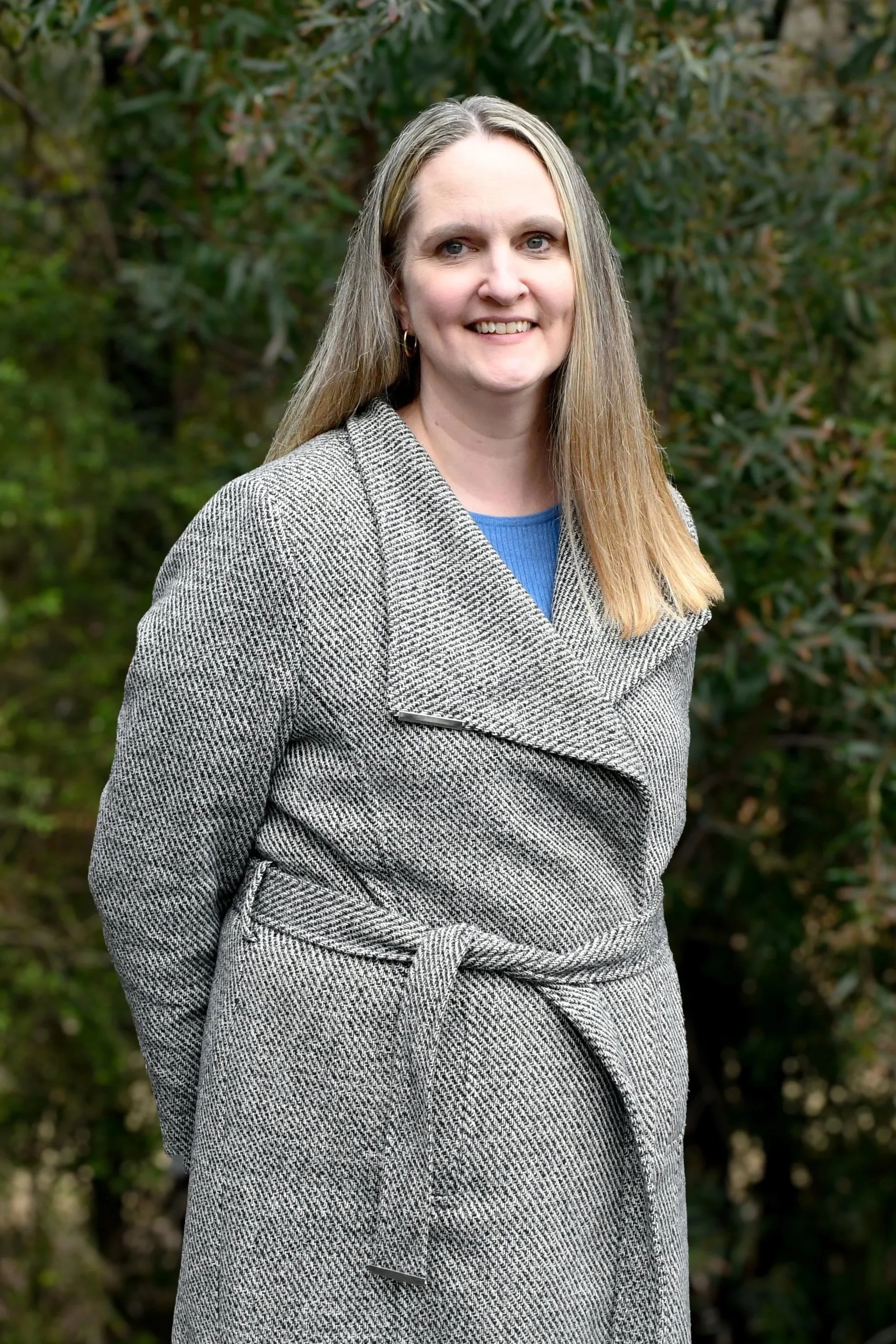 Woman with chest length ash blonde hair wears a wrap style coat over a blue top, she is smiling at the camera.