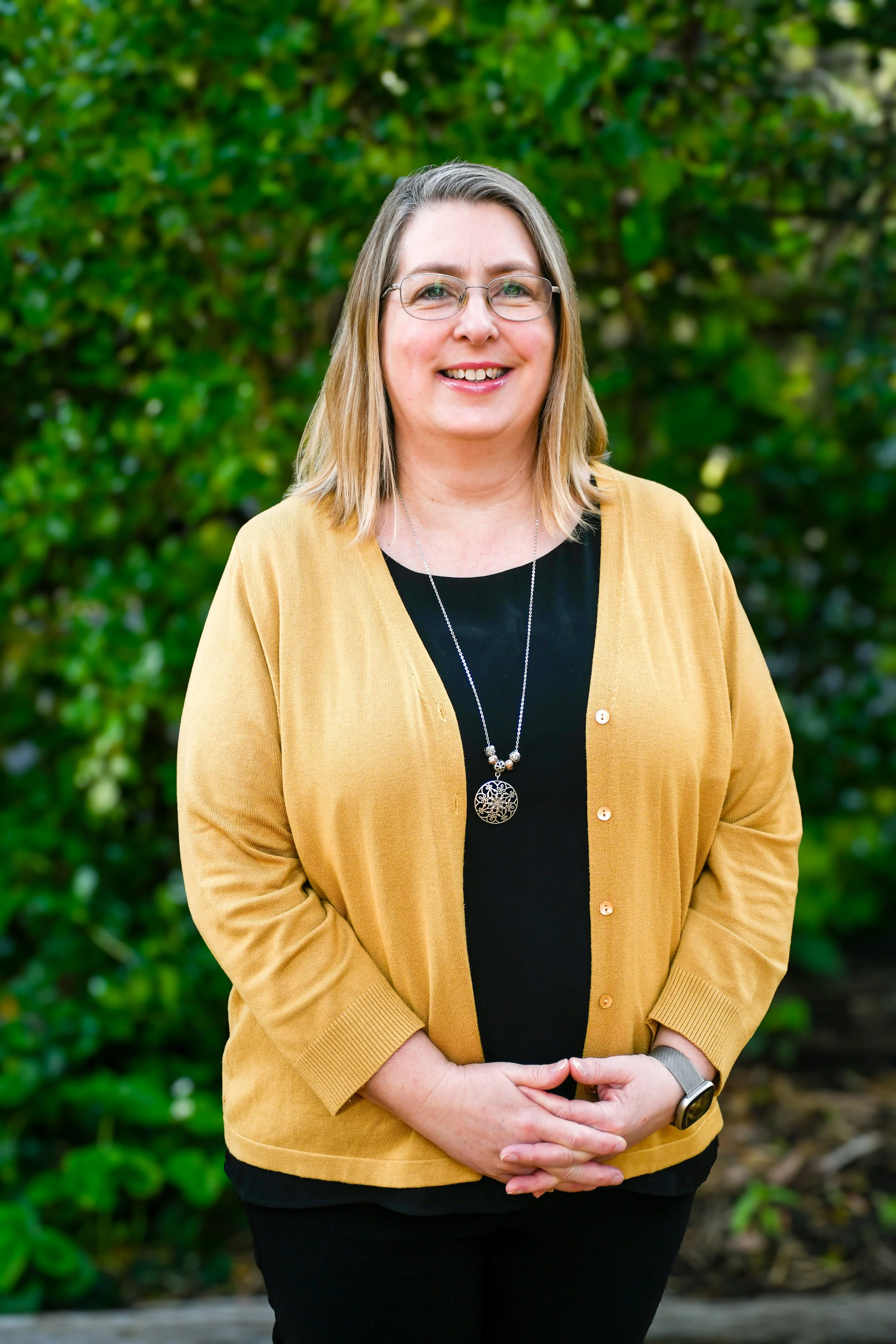 A woman is wearing a mustard yellow cardigan over a black t-shirt. She has shoulder length brown hair with blonde highlights and wears glasses with a wire frame. She is smiling at the camera.