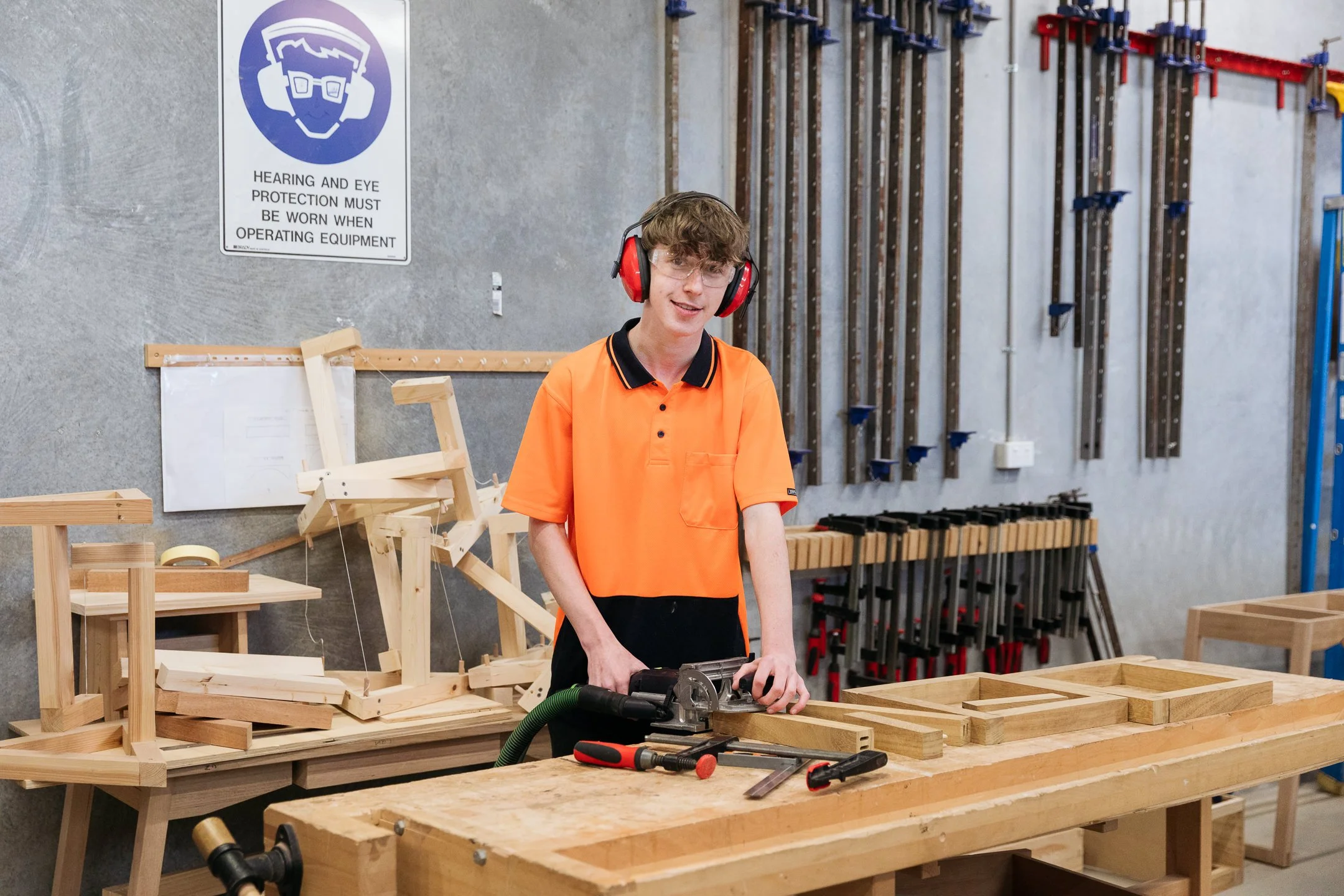 Male high school student wearing orange high-vis polo shirt and protective ear muffs and eye-wear works at a construction work bench