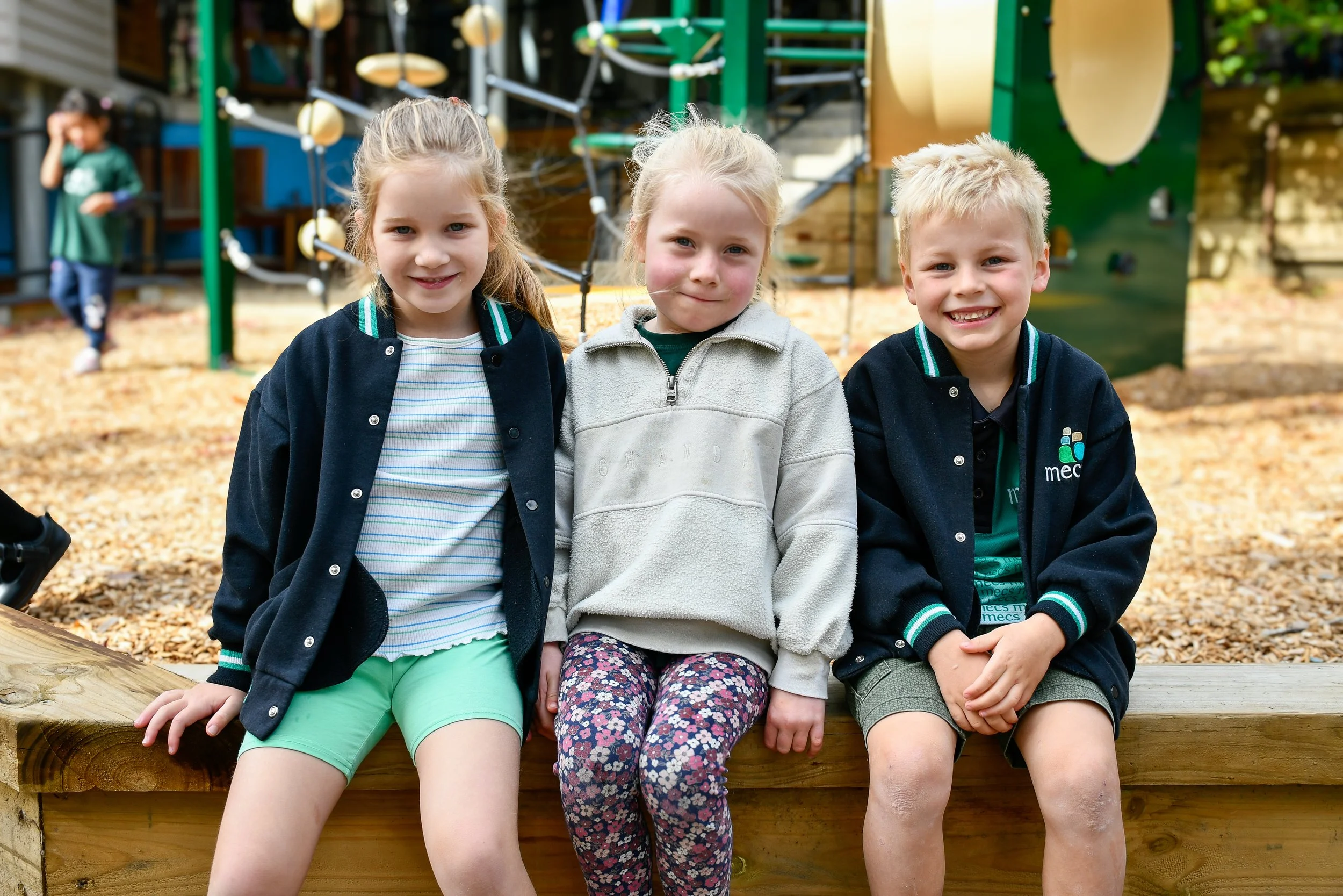 3 junior primary students seated in playground smiling at the camera. There are two girls on left and middle and one boy on the right, they all have light blonde hair and are in free dress.