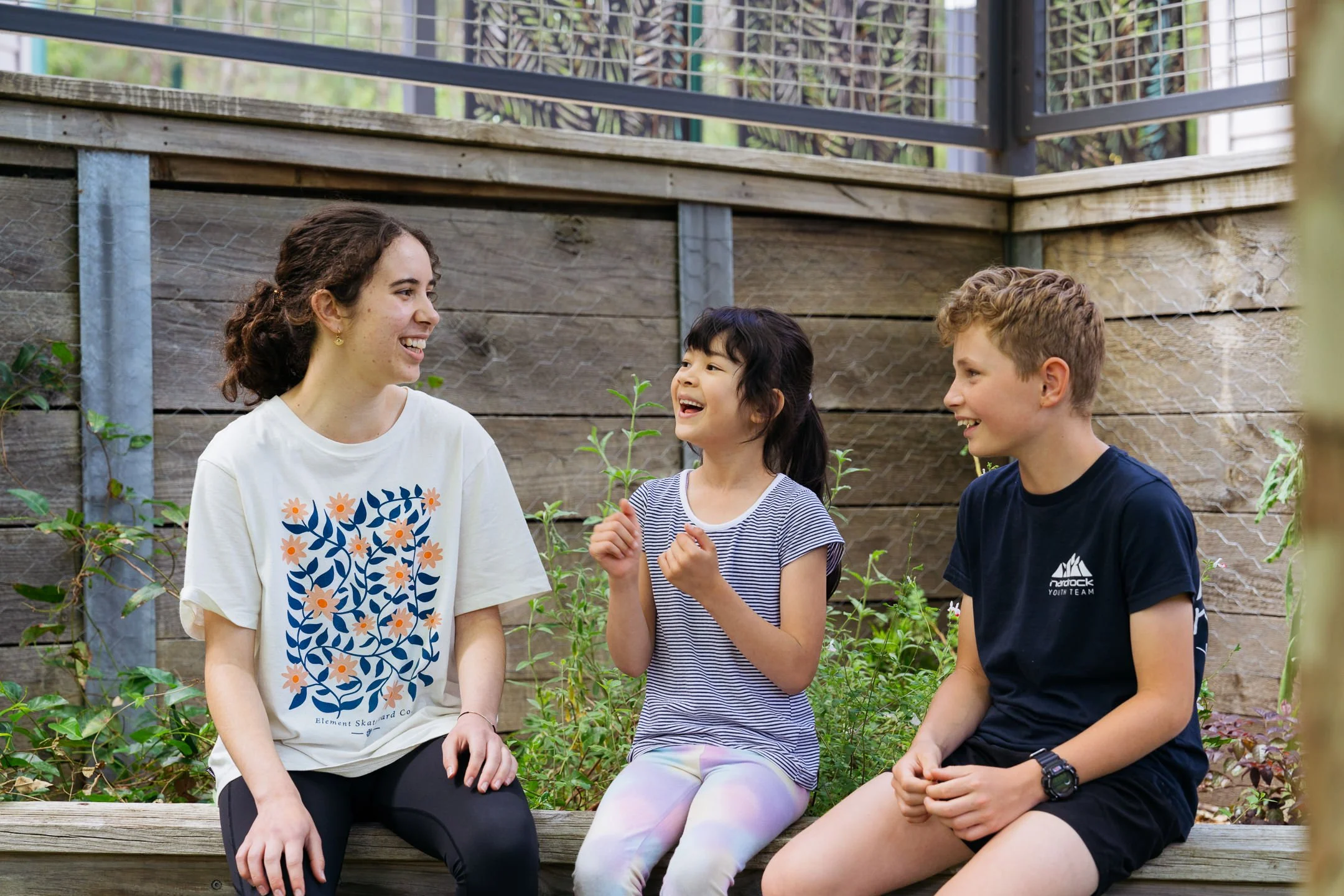 Two young children sitting outdoors on a wooden bench with an older student, laughing and talking with each other in a garden with greenery and a wooden fence in the background.