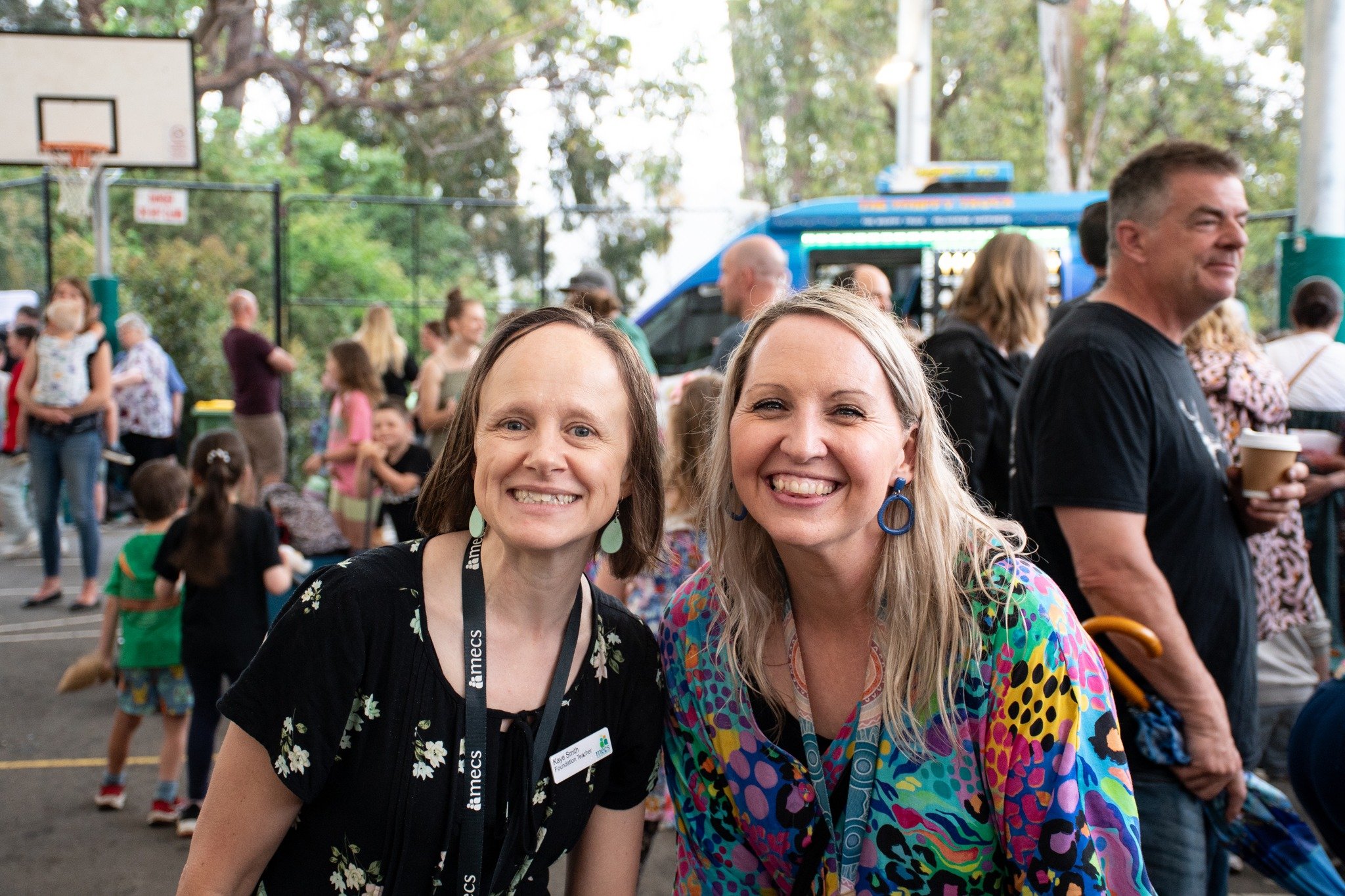 Two female teachers smiling at camera in front of a crowd of people mingling at a carnival