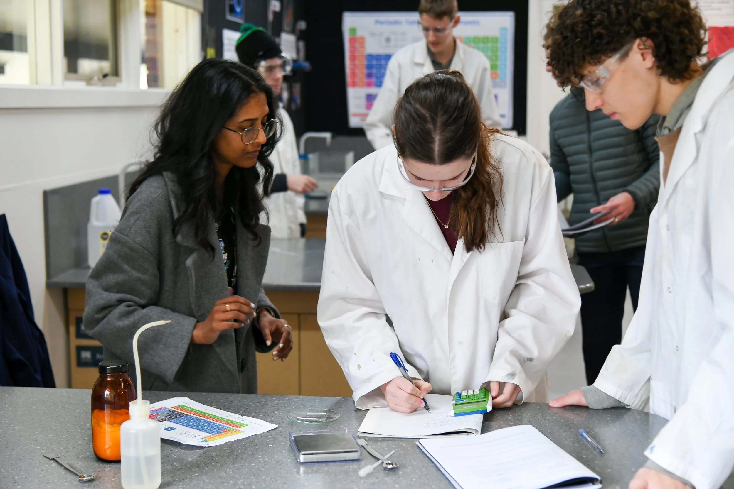 Science teacher oversees the work of two students wearing lab coats and protective eye wear