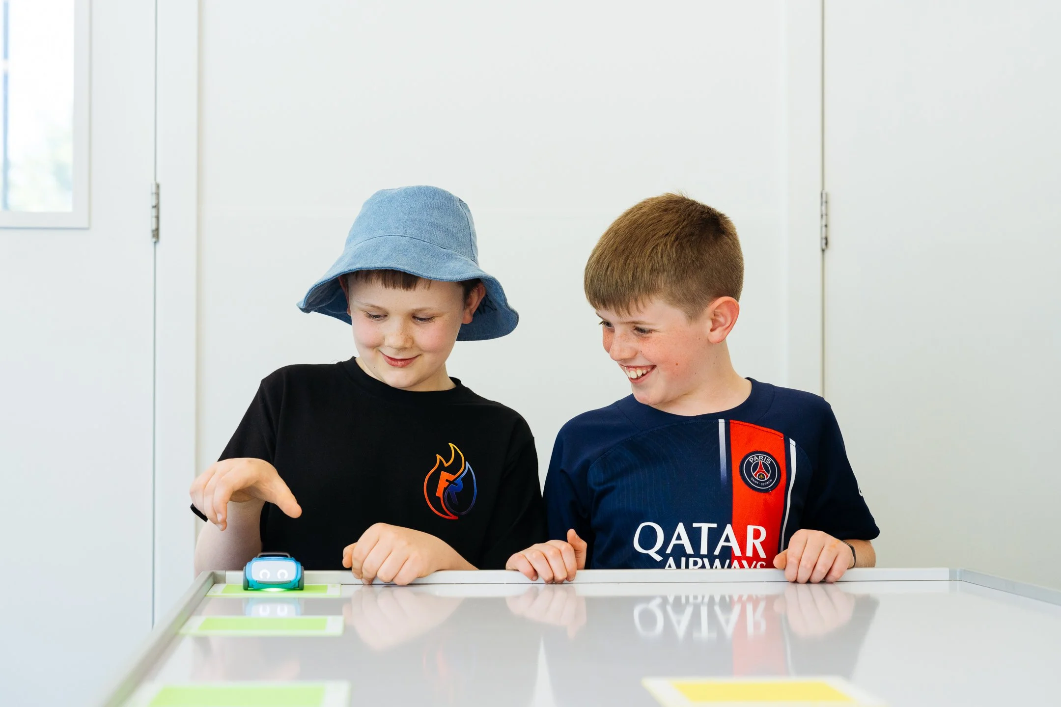 Two male Senior Primary students are working at a desk. They are smiling, looking down at the robotic device on the table.