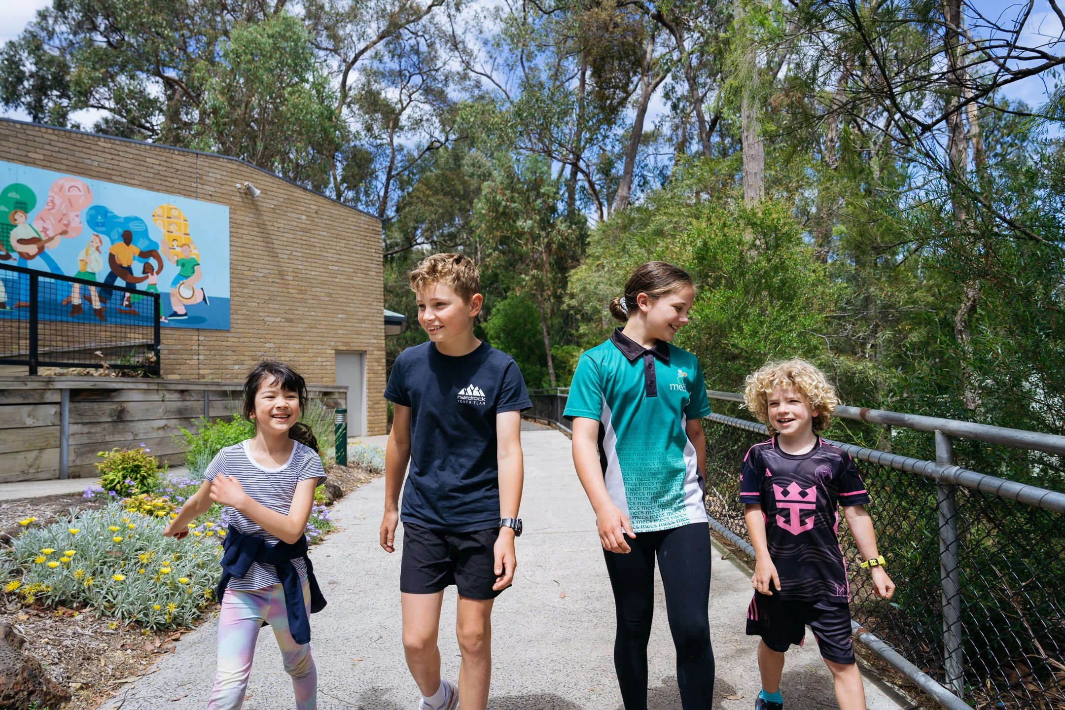 4 students casually walking down pathway. From left to right, there is a young Junior Primary female student, an older Senior Primary male student, an older Senior Primary female student and then a young Junior Primary male student.