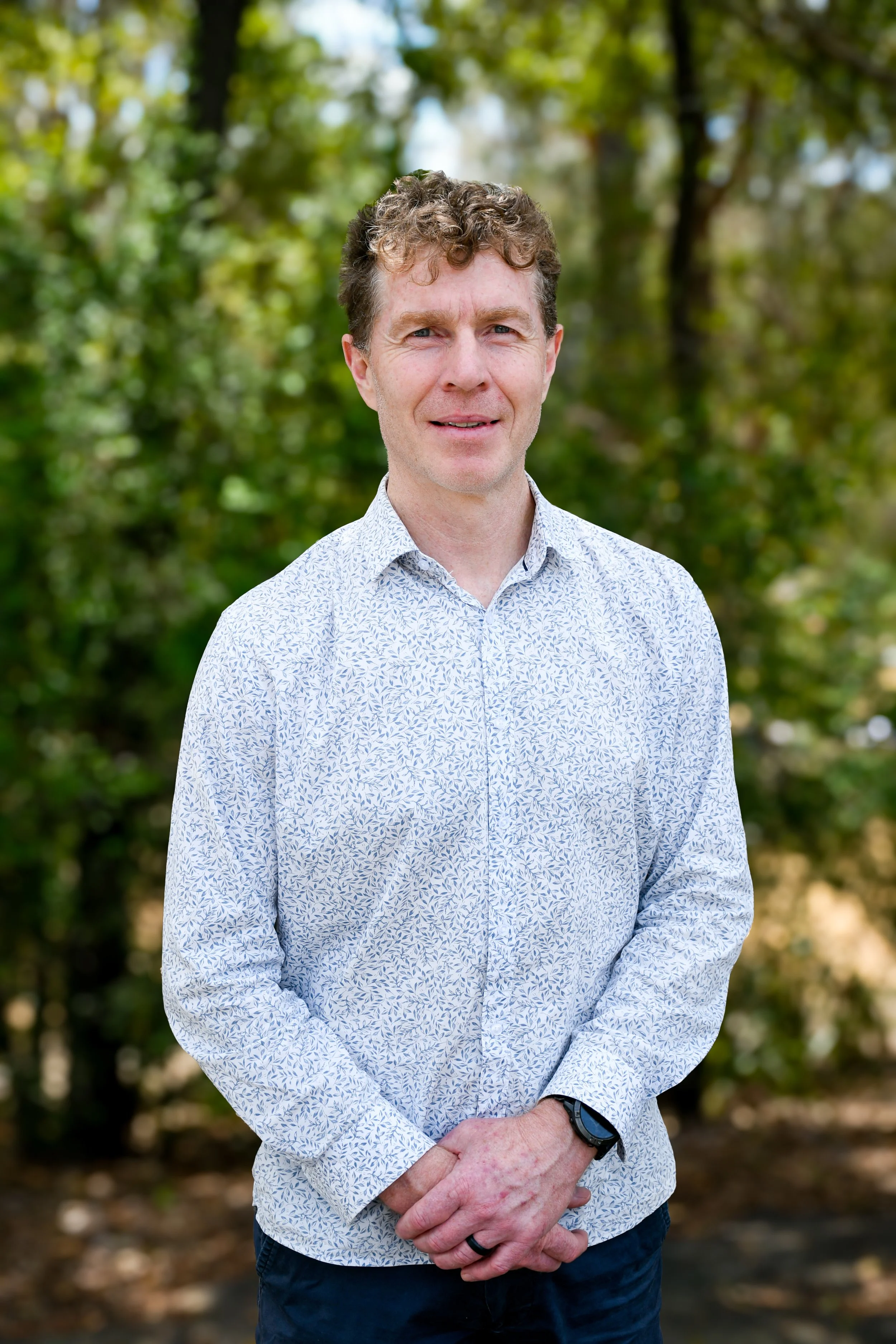 A man wearing a white & grey patterned button up shirt. He has short, brown hair and is smiling at the camera.