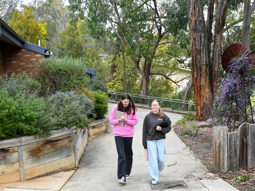 Two female high school aged students walking and talking outdoors on a sidewalk, one holding a notebook and the other with a binder, surrounded by trees and plants.