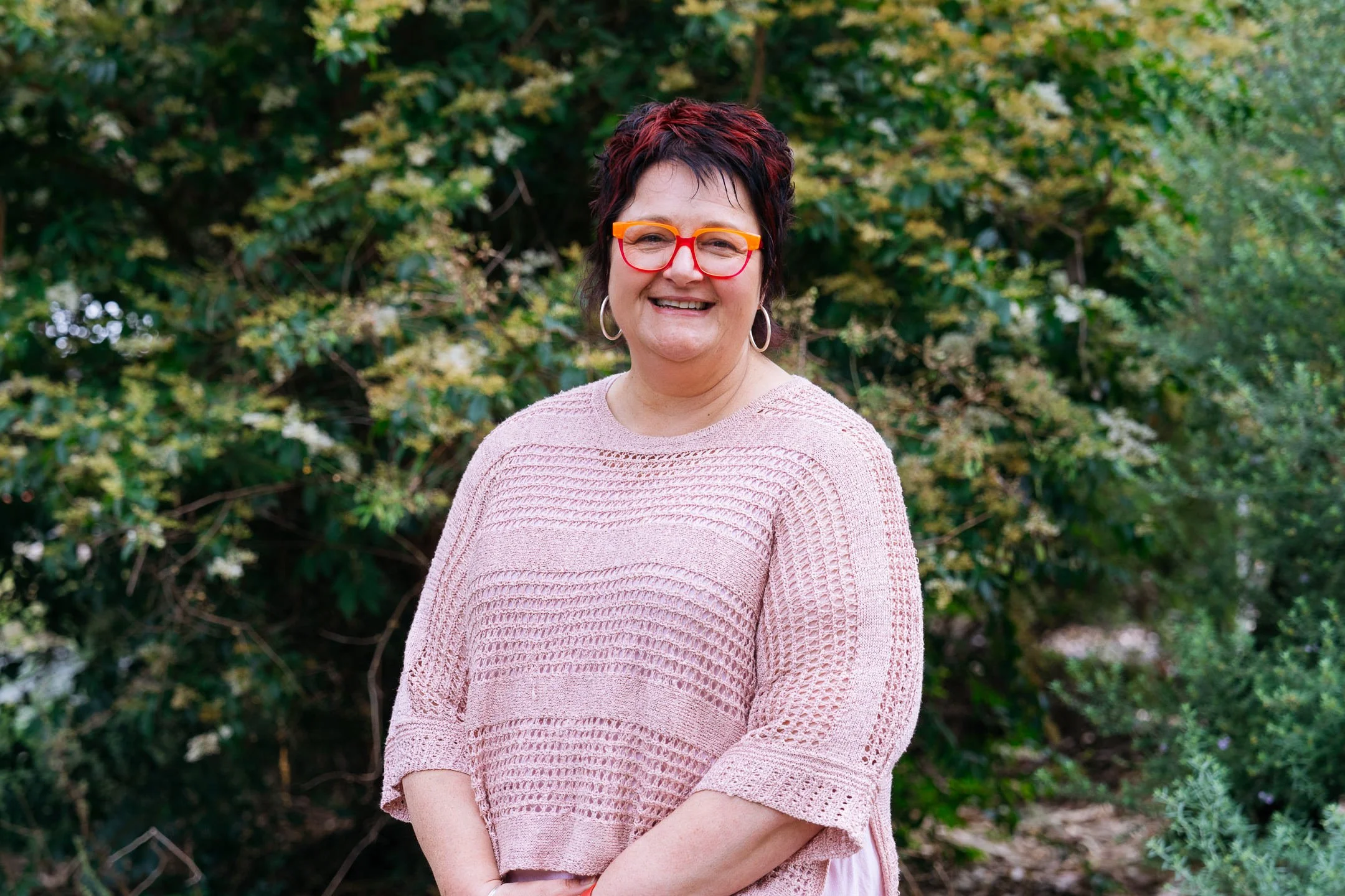 Principal, Michelle Dempsey, standing in front of green hedge, wearing a light pink top with pink and orange glasses on