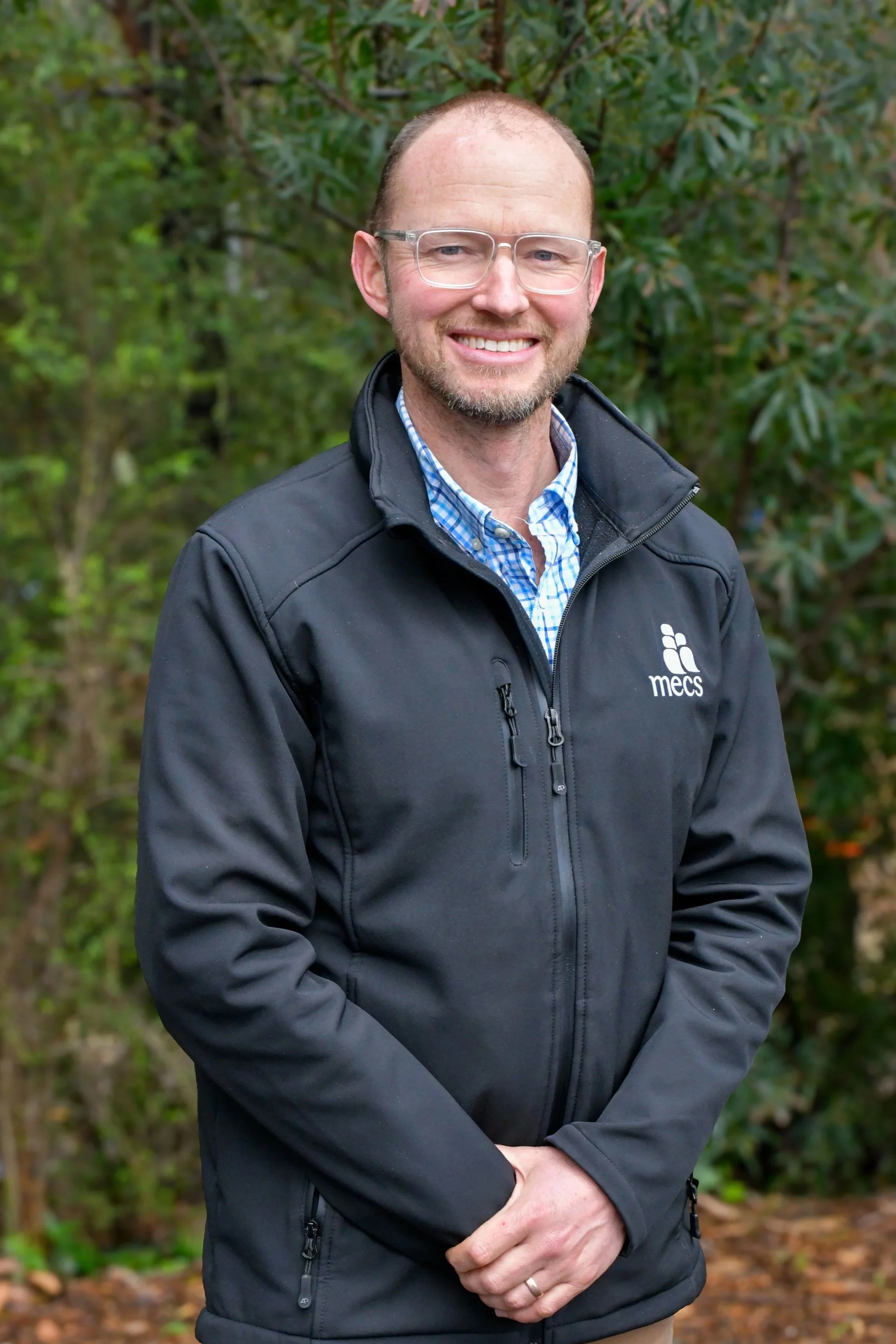 A man wears a black jacket with the MECS school logo on the breast pocket over a checkered blue formal shirt, he wears transparent framed glasses and is smiling at the camera.