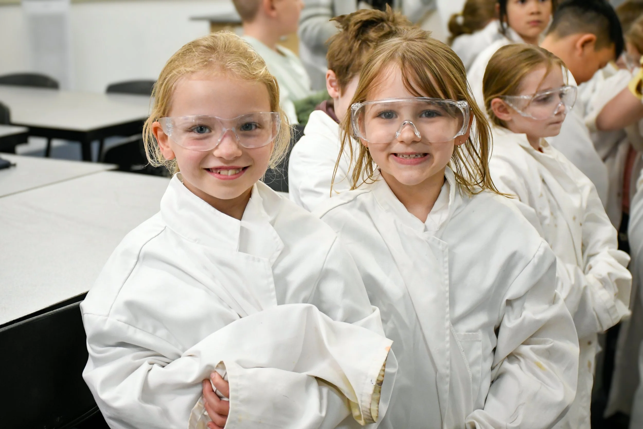 Two female Prep-aged students are wearing white lab coats and protective eyewear, smiling at the camera.