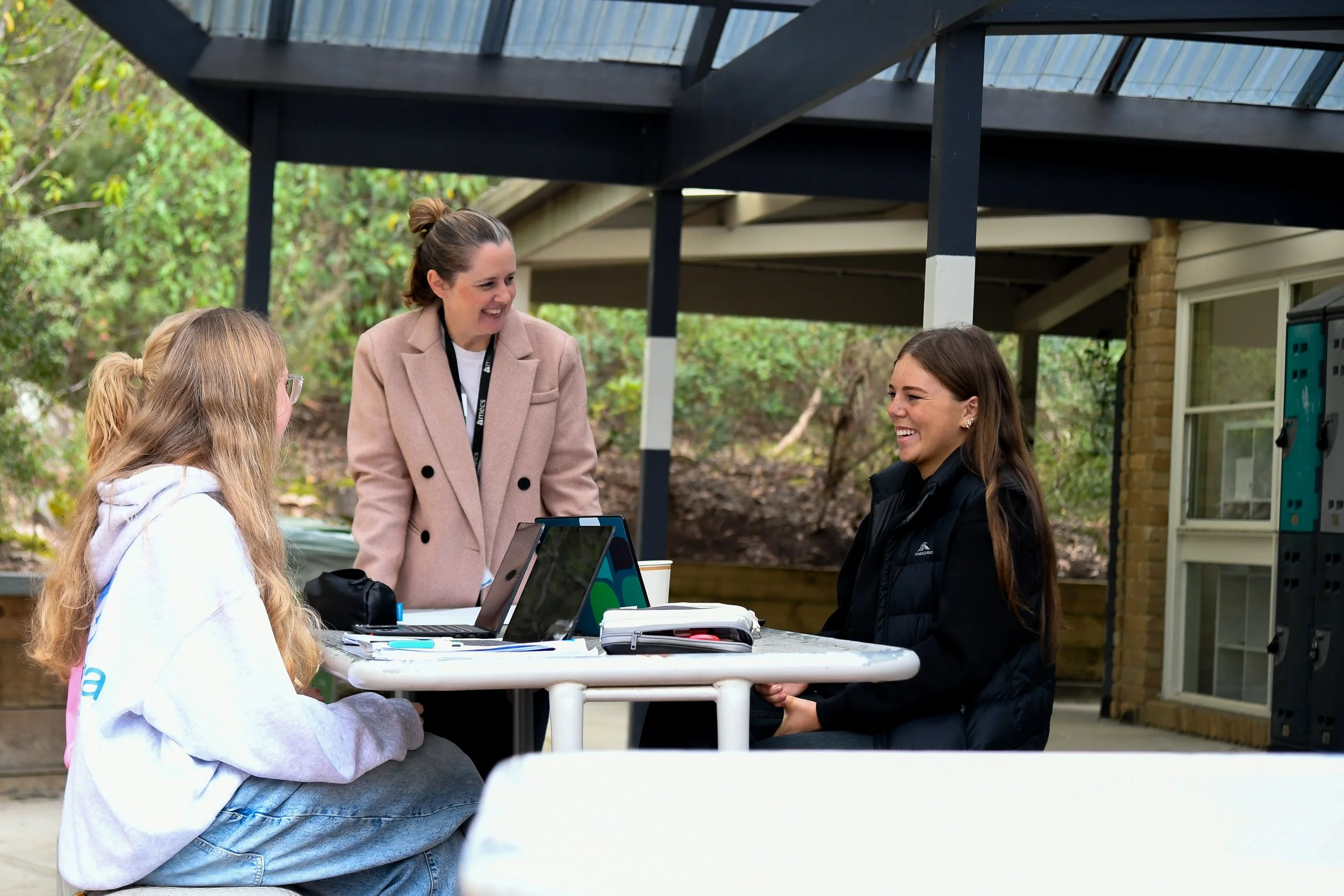 3 females gathered around picnic table, two students are seated and the teacher is standing