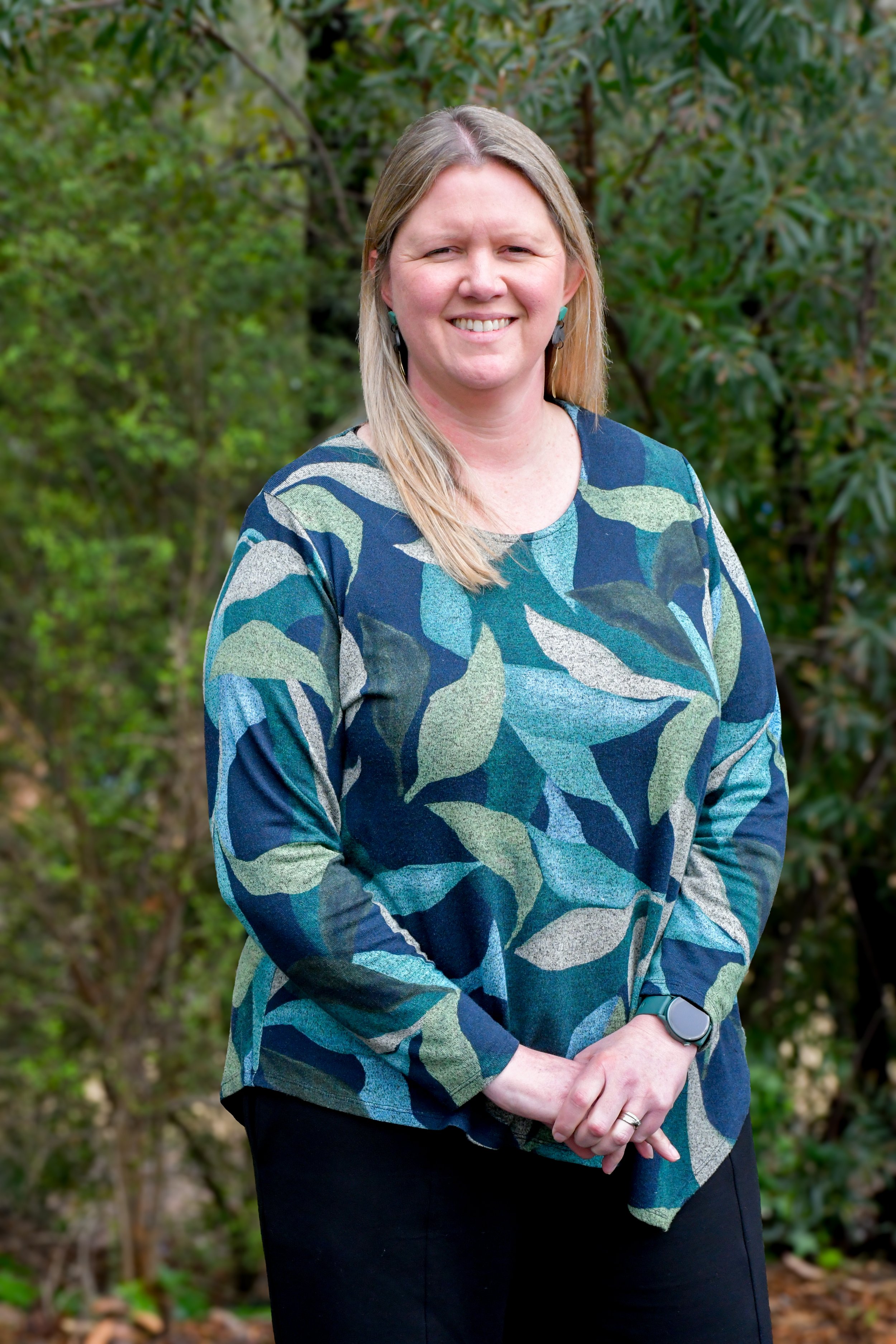 A woman wearing a blue & green leaf patterned long sleeve top. She has long blonde/ brown hair that is worn down and she is smiling at the camera.