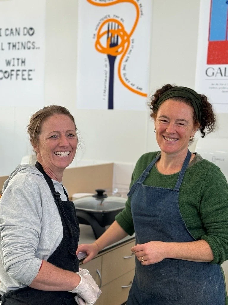 Two adult women working in kitchen, wearing aprons and smiling at the camera.