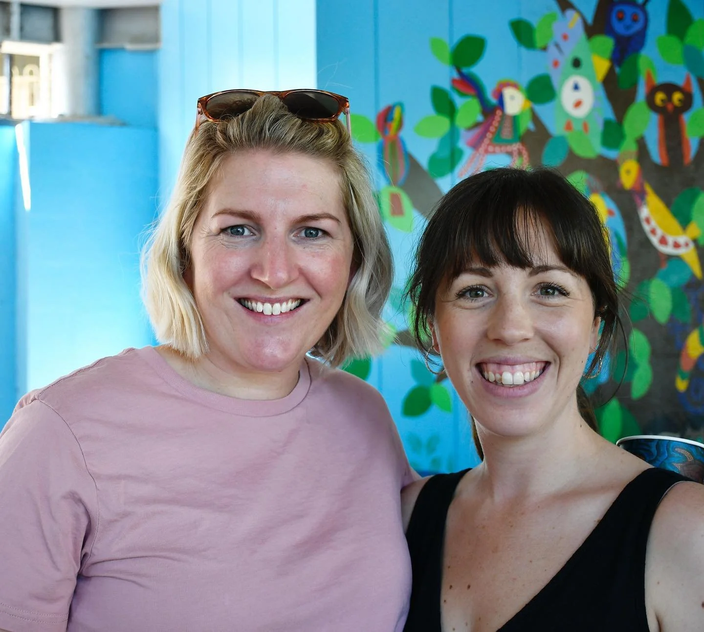 Two adult women smiling at the camera. On the left, the woman has a short blonde bob with sunglasses on top of her head and a light pink t-shirt. On the right, the women has dark brown hair with a fringe and is wearing a black singlet.