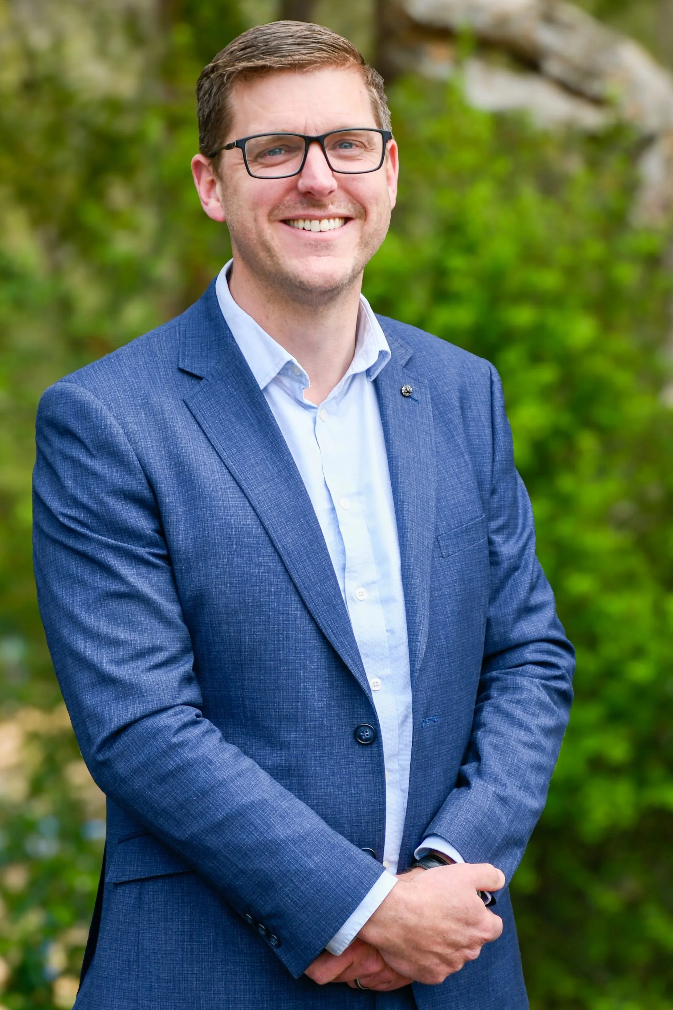 A man is a wearing a blue blazer over a light blue formal shirt. He has short, brown hair and black rectangular glasses. He is smiling at the camera.