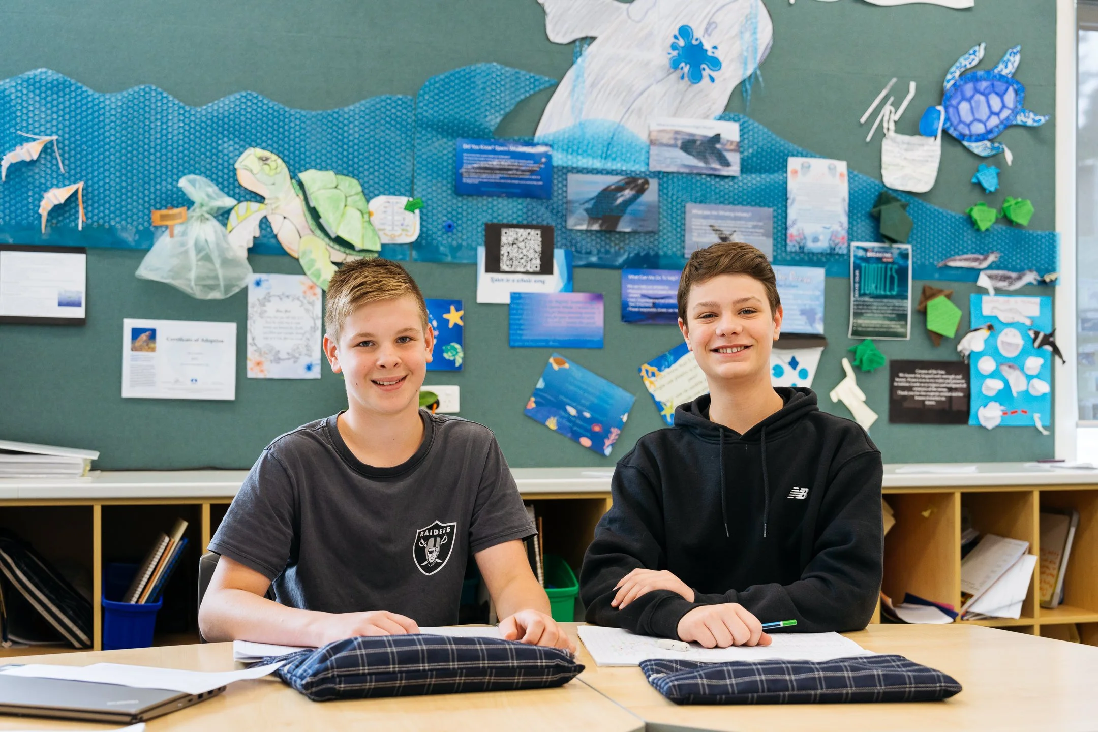Two boys sitting at a school desk in front of a colorful ocean-themed bulletin board with marine creatures and informational posters, smiling at the camera.