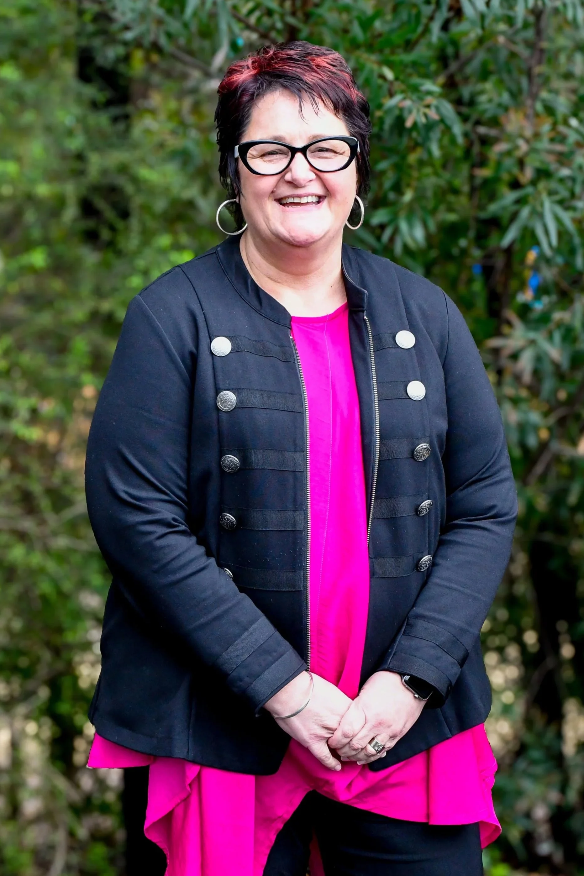 Woman with a black jacket on, seen underneath is a hot pink blouse. She is wearing bold black oval glasses and has a short pixie cut hairstyle that is black with red highlights. She is smiling at the camera.