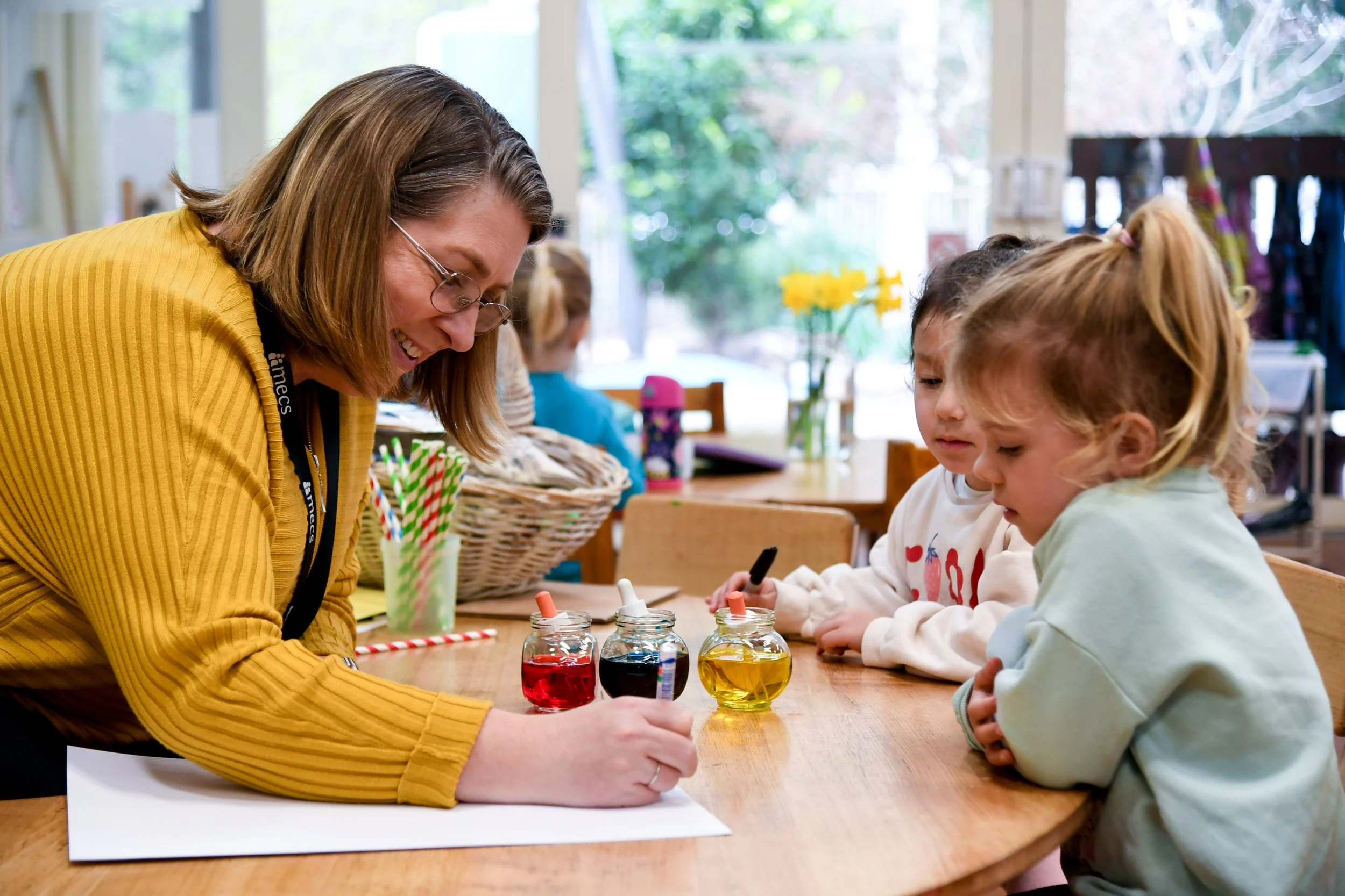 Kindergarten teacher interacts with two pre-school aged students, creating a craft at a table together