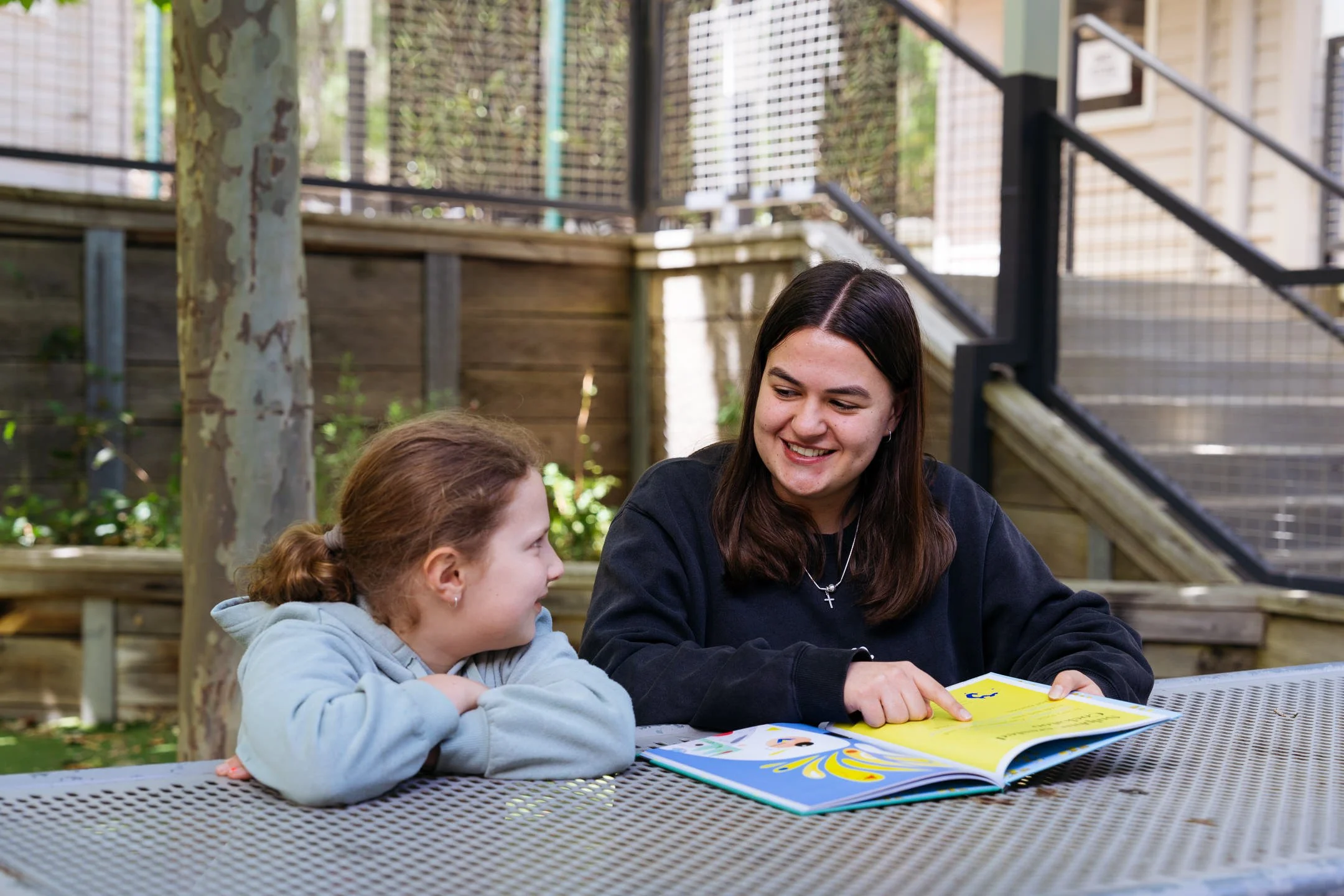 A senior female student and a young girl sitting at a metal table outdoors, looking at a colourful children's book and smiling.