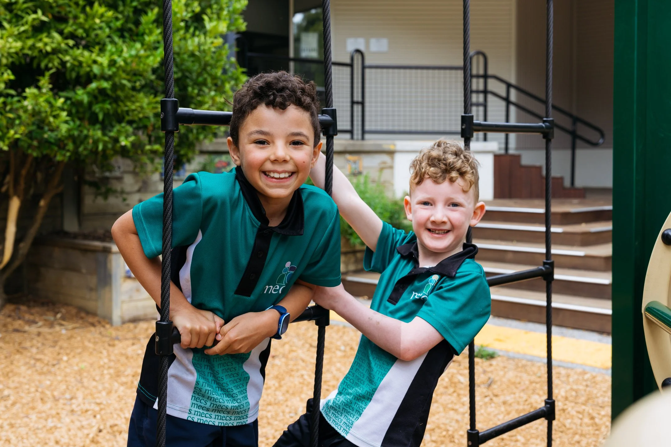 Two male junior primary school aged boys climb on playground equipment. They are wearing school polos and smiling at the camera.