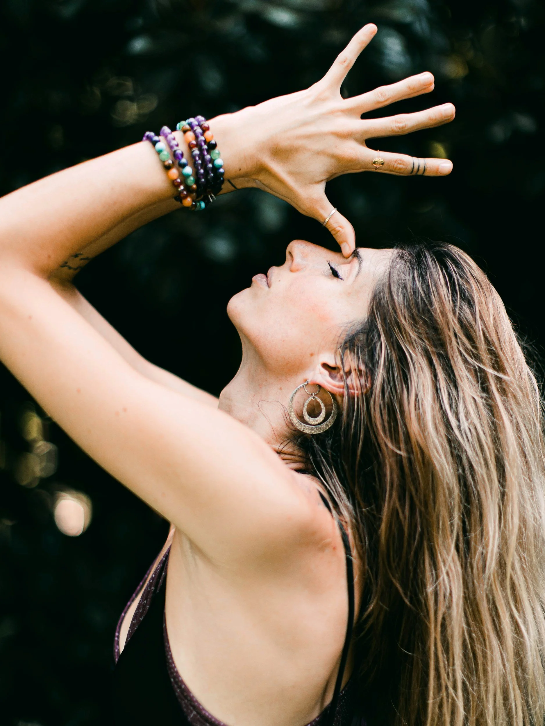 A woman with long, wavy hair and earrings poses with her eyes closed, touching her forehead with her fingers in a graceful pose. She wears multiple colorful beaded bracelets and rings, with a dark background.