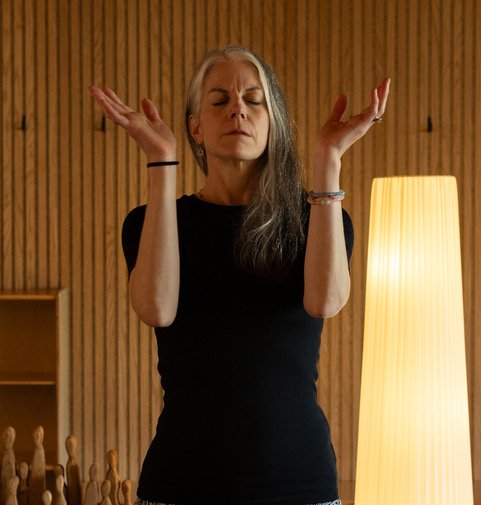 A person practicing yoga or meditation in a seated lotus position, with one hand resting on the knee and the thumb and index finger touching in a mudra, wearing a pink top and blue leggings, sitting on a wooden floor.