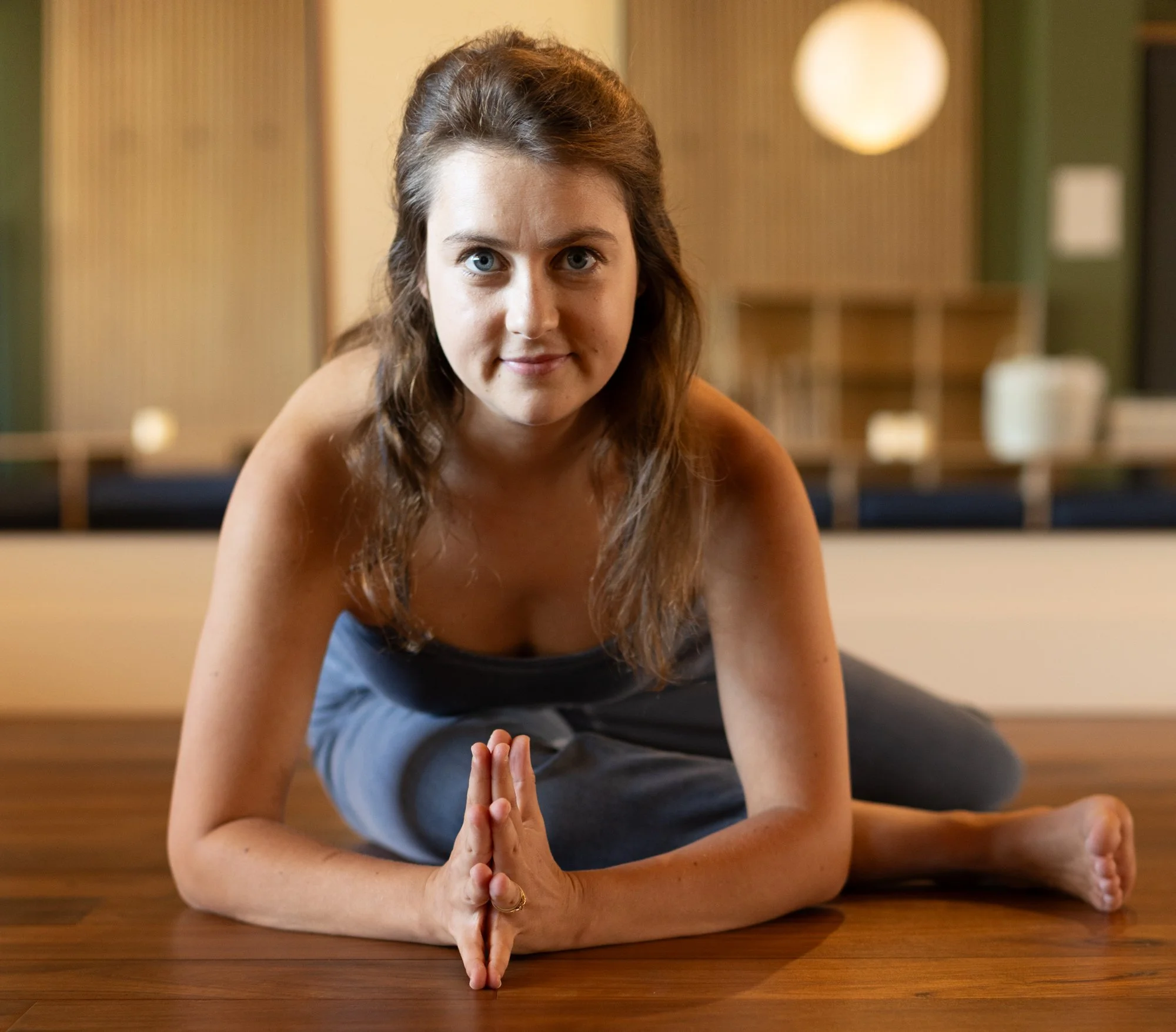 A person with long dark hair practicing yoga in a child's pose on a red mat indoors near a window with curtains, with trees visible outside.