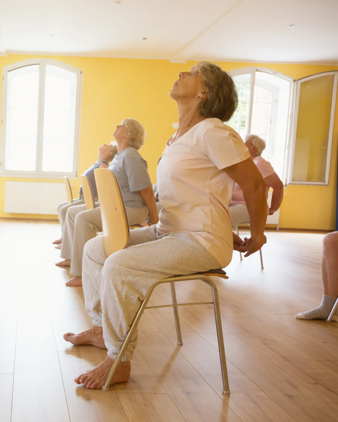 Older women participating in a seated stretching or yoga class in a bright room with yellow walls and large windows.