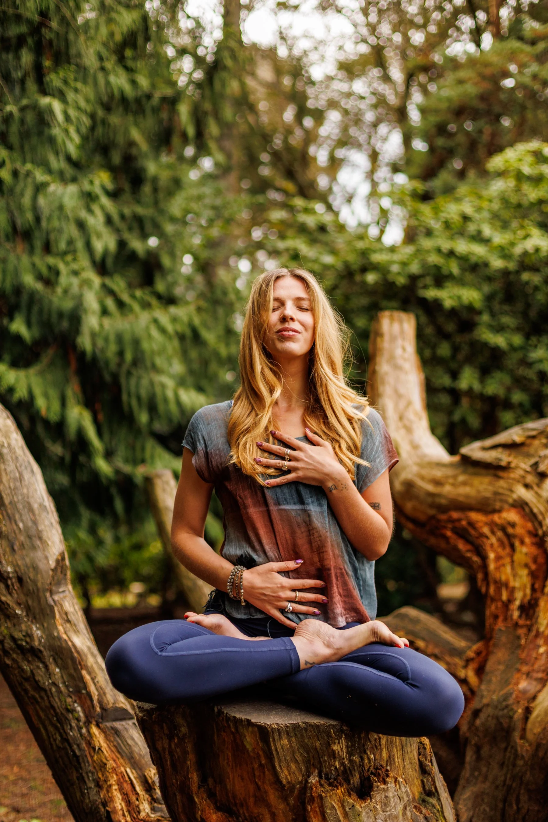 A woman practicing yoga outdoors on a tree stump in a forest, sitting cross-legged with hands on her chest and stomach, eyes closed, appearing peaceful and relaxed.