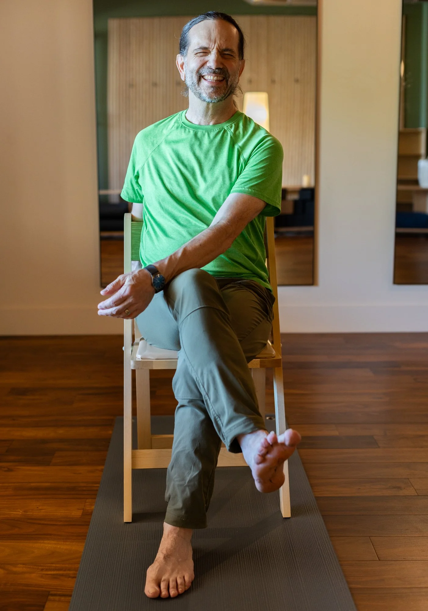 Older women participating in a seated stretching or yoga class in a bright room with yellow walls and large windows.