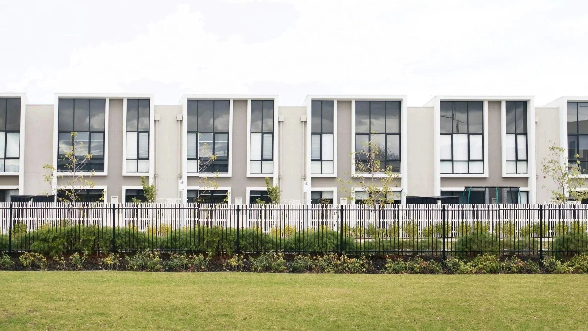 Modern multi-story residential building with large windows, a black metal fence, and landscaped yard with trees and shrubs.