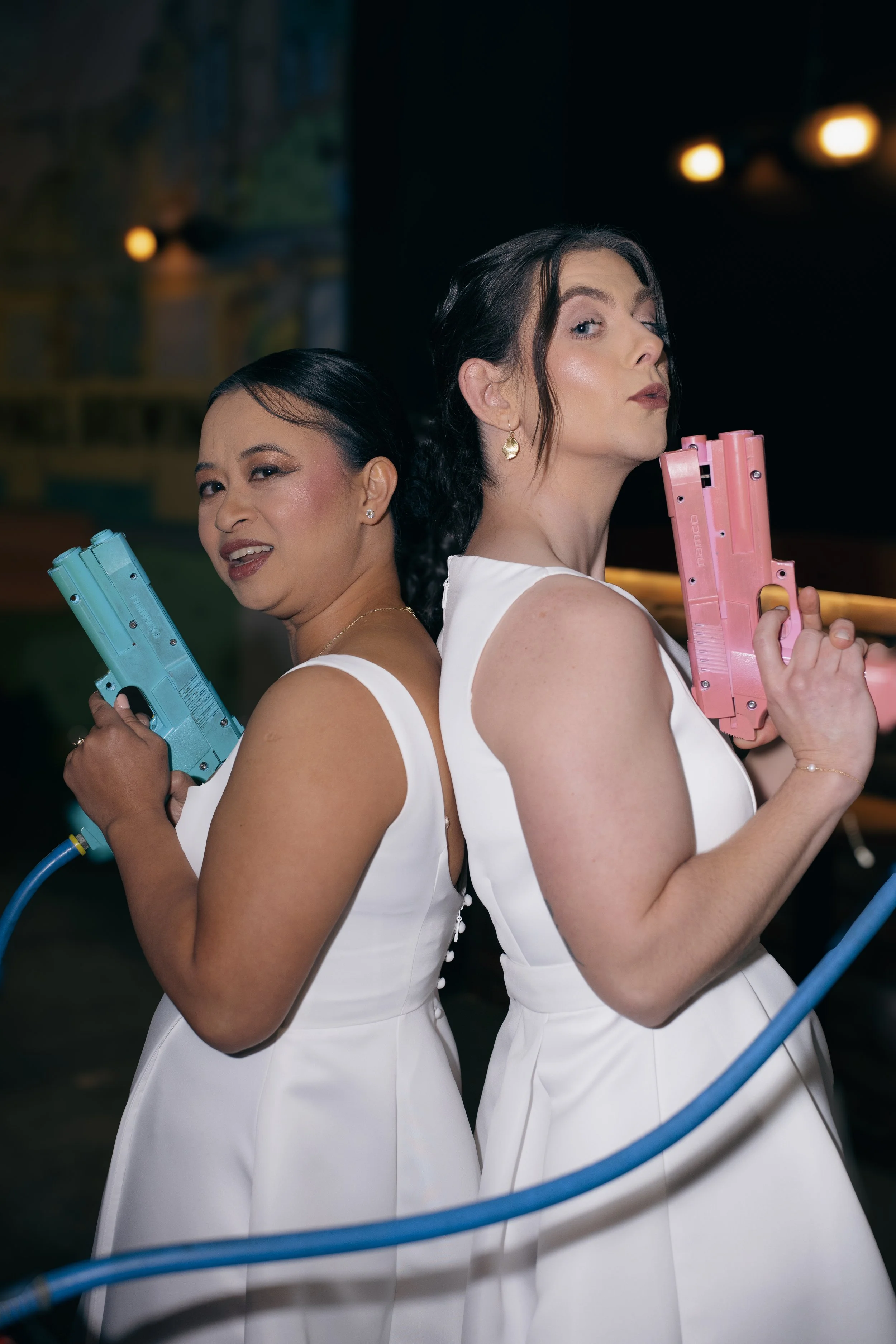Two women in white dresses holding toy water guns back to back, posing confidently in a dimly lit indoor setting.