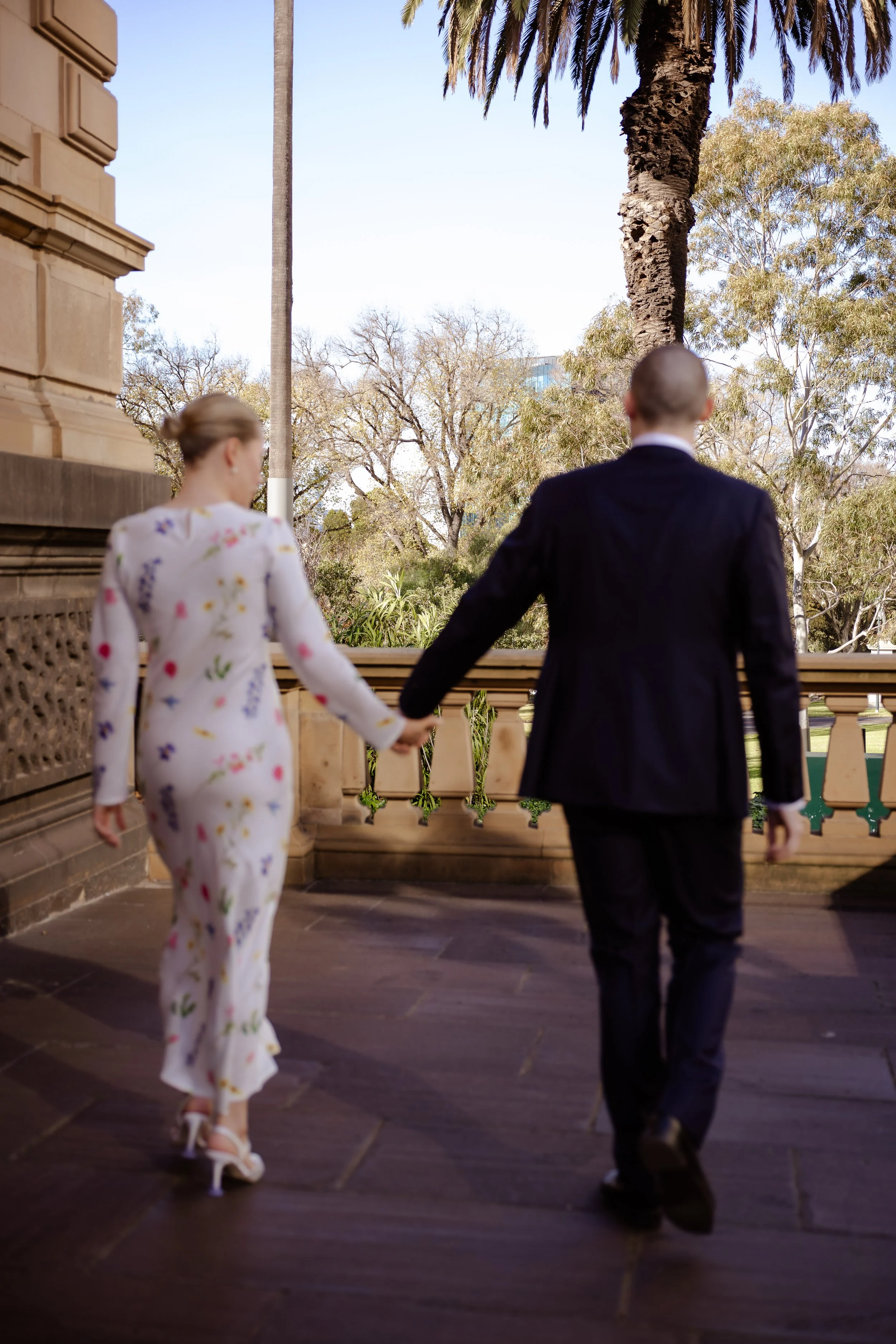 A couple walking hand in hand on a balcony outside a building, with a decorative railing. The woman is wearing a white floral jumpsuit and shoes, while the man is dressed in a dark suit. Behind them are trees and a clear sky.