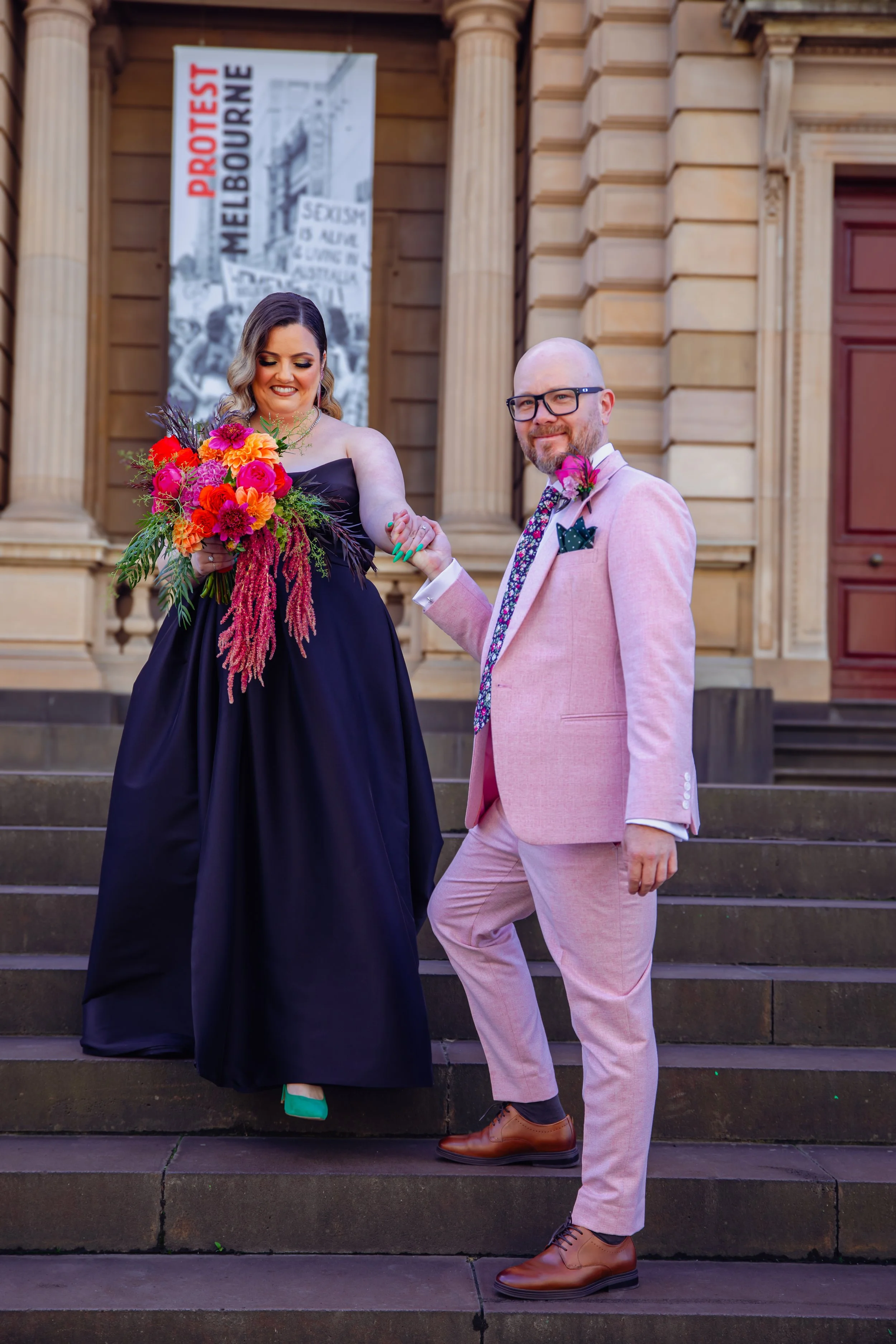 A couple standing on steps outside a historic building, holding hands. The woman is wearing a black dress and holding a colorful bouquet of flowers. The man is dressed in a light pink suit with a floral shirt and brown shoes. There is a protest banne
