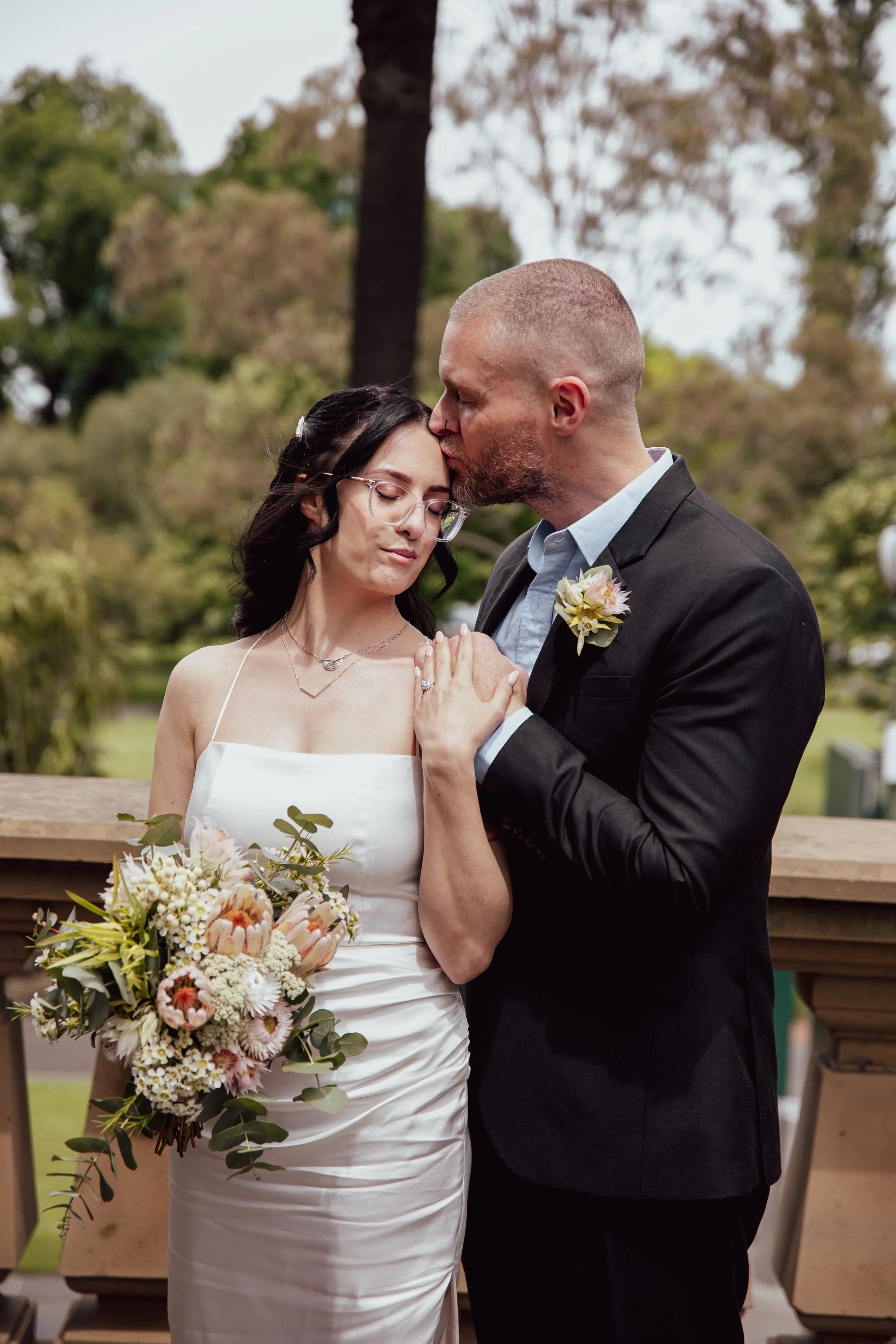 A bride and groom share a tender moment during their wedding, with the groom kissing the bride's forehead as she closes her eyes, holding a bouquet of flowers, standing outdoors in front of a park.