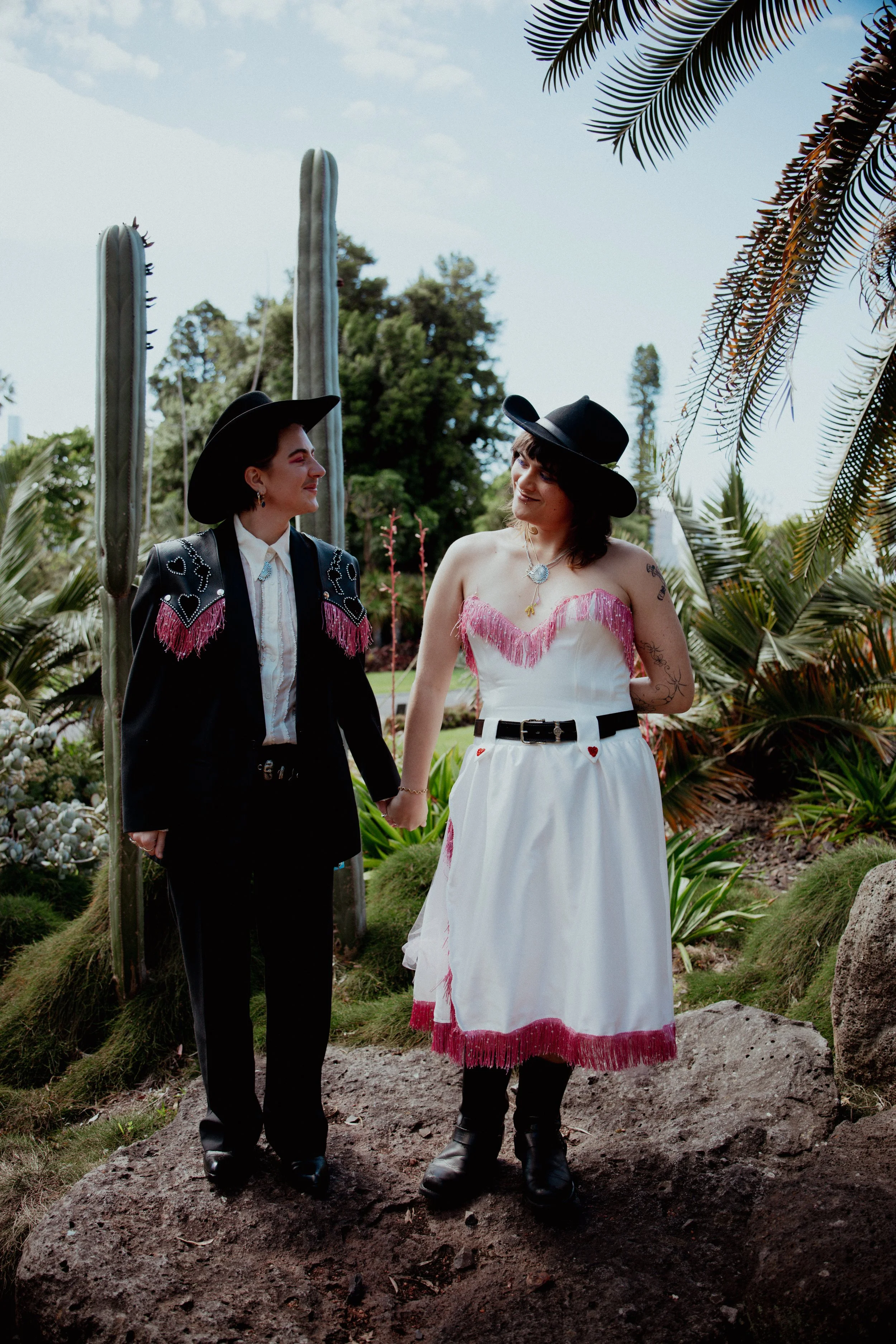 Two women holding hands in a garden with cacti and lush plants, wearing black hats and eccentric clothing.