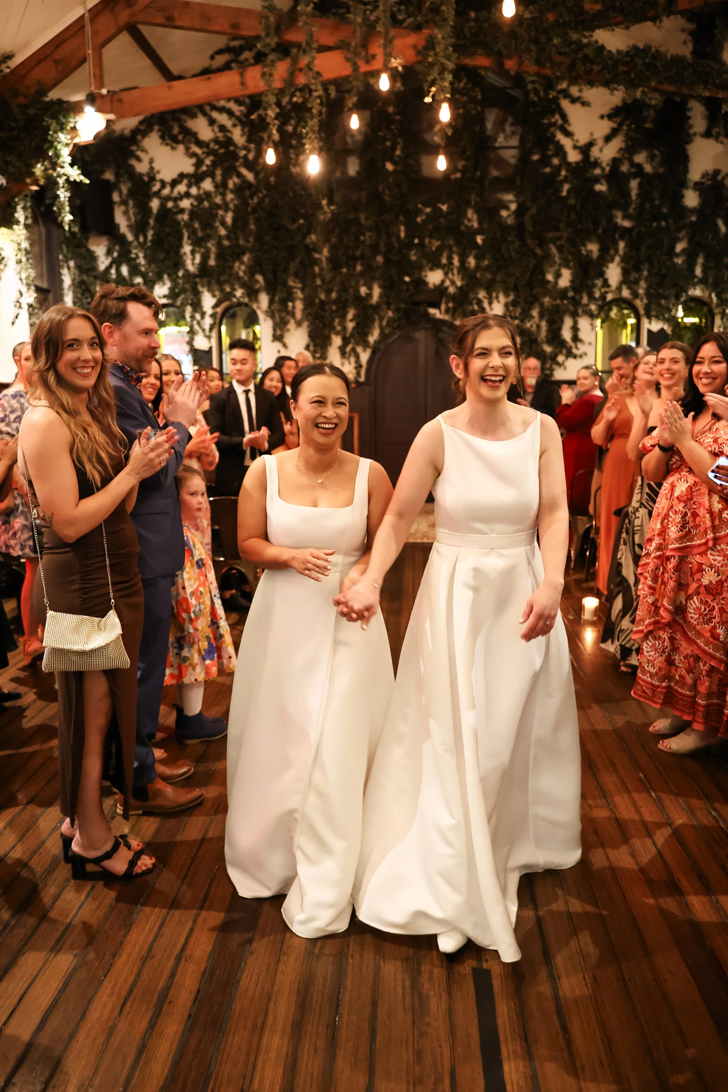 Two women in white dresses holding hands and smiling, surrounded by happy people at a wedding reception with wooden floors and decorated ceiling.