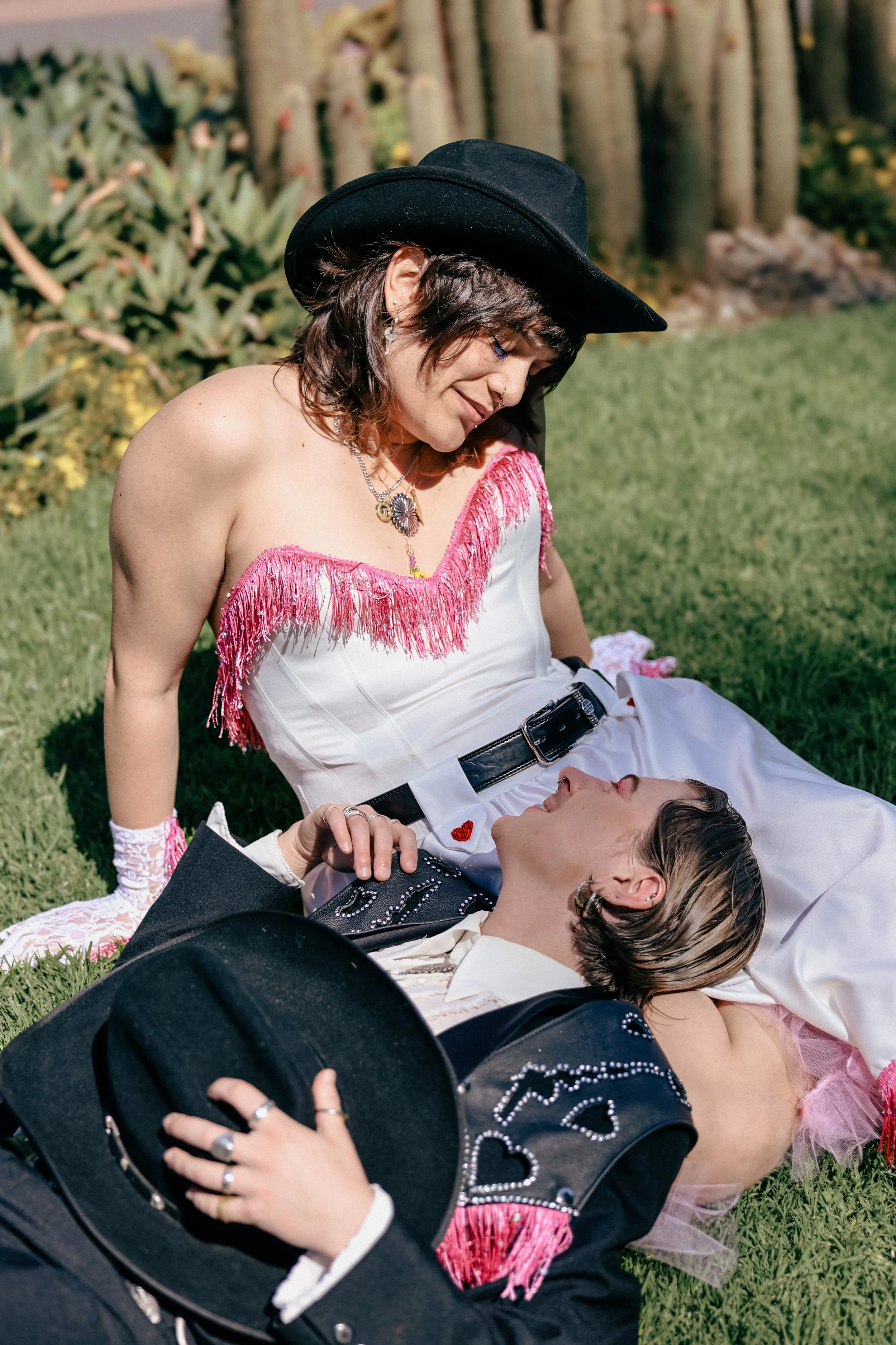 Two women dressed in western attire, one sitting on the grass and the other lying down, sharing an affectionate moment outdoors with greenery and trees in the background.