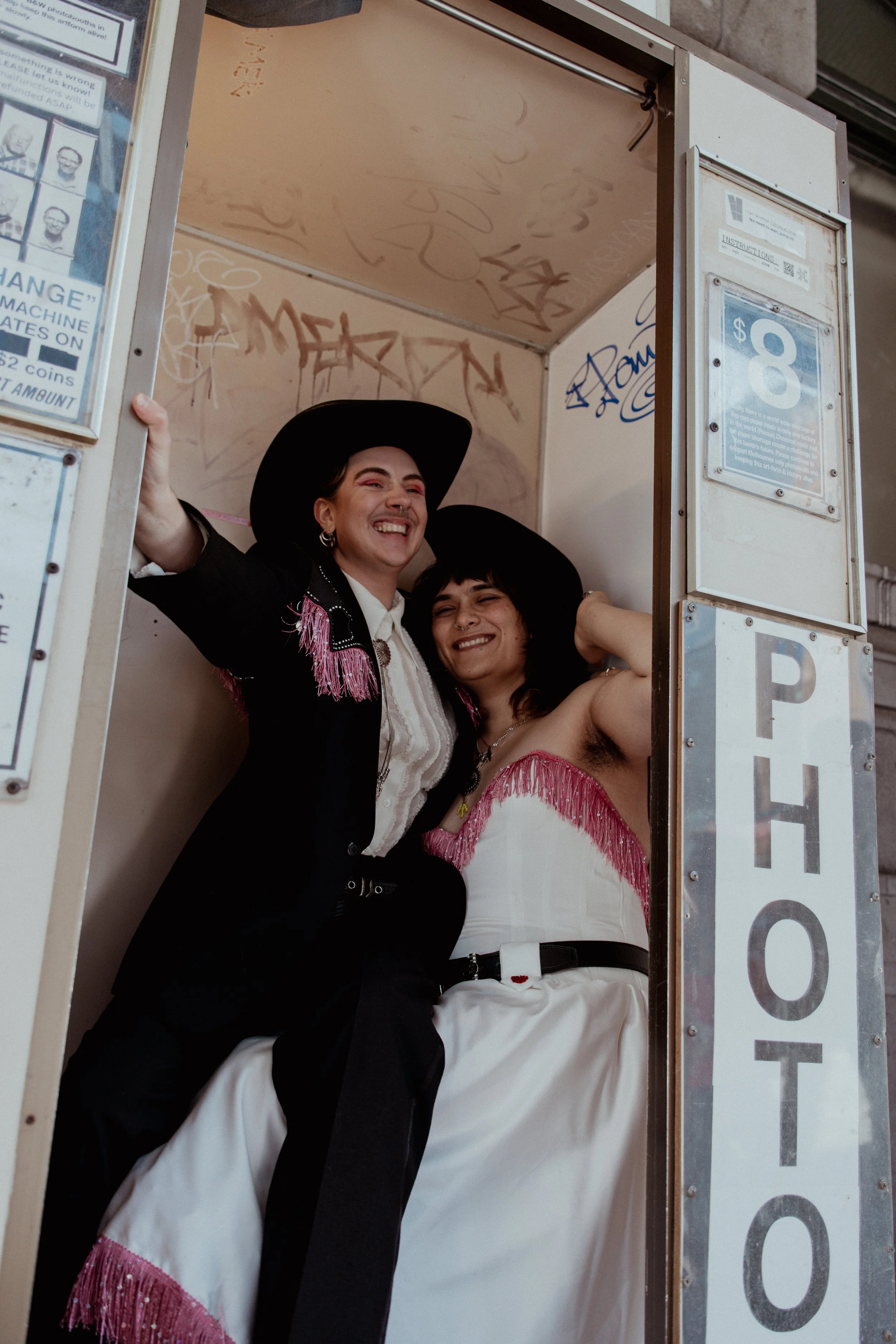 Two women wearing western-themed outfits and large black hats, smiling and laughing inside a small photo booth.