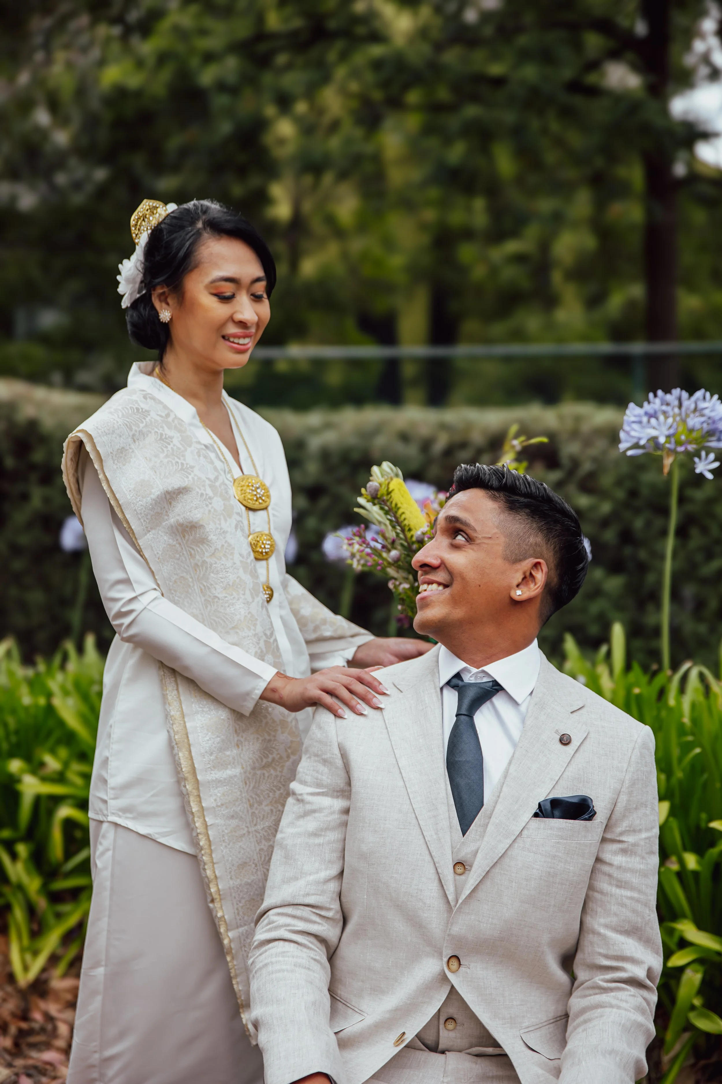 A couple dressed in formal attire, the woman in traditional Asian clothing and the man in a light-colored suit, sharing a moment outdoors with greenery and flowers in the background.